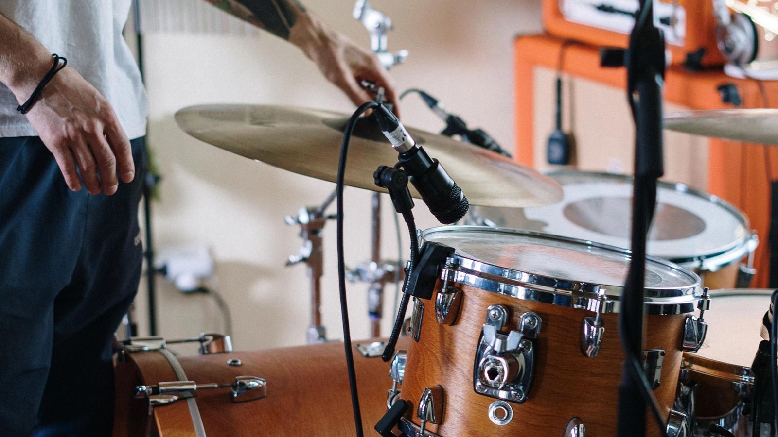 A drummer adjusts a cymbal microphone in a home rehearsal room, with a wooden drum kit in the foreground and guitar amps against the wall behind.