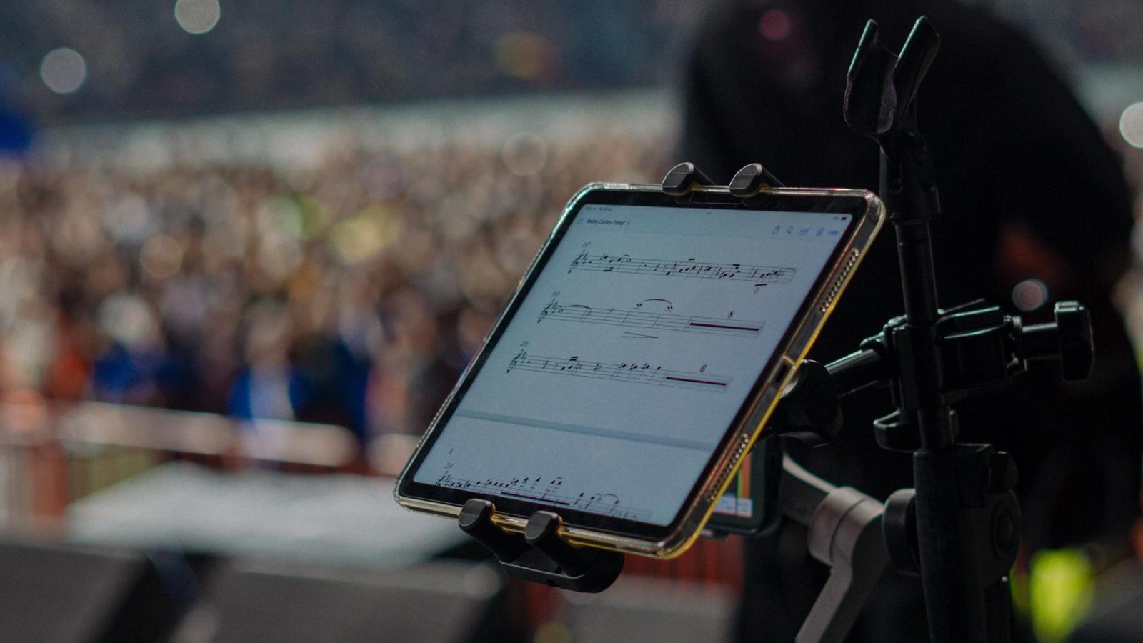 A tablet mounted on a mic stand displays sheet music on stage, with a guitarist and a stadium crowd blurred behind it and camera gear in the foreground.