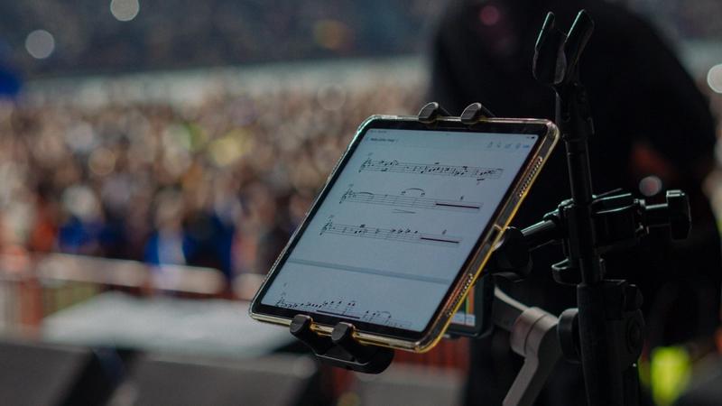 A tablet mounted on a mic stand displays sheet music on stage, with a guitarist and a stadium crowd blurred behind it and camera gear in the foreground.