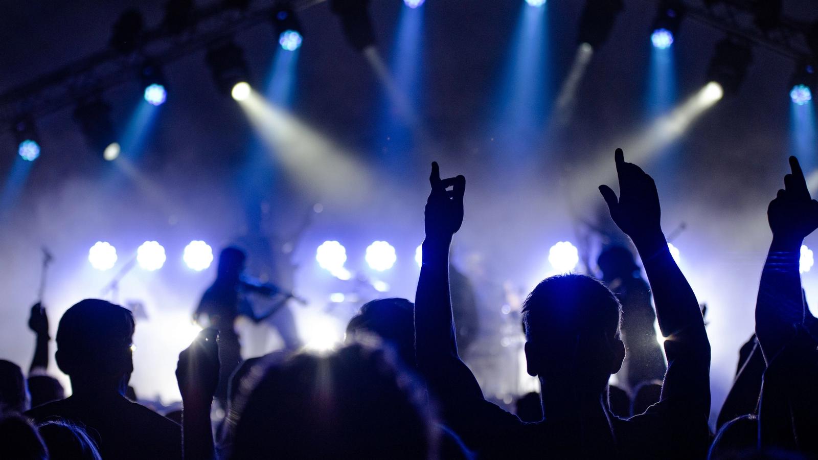 A crowd with hands raised in front of a stage lit by blue and white lights, with a band silhouetted behind them.