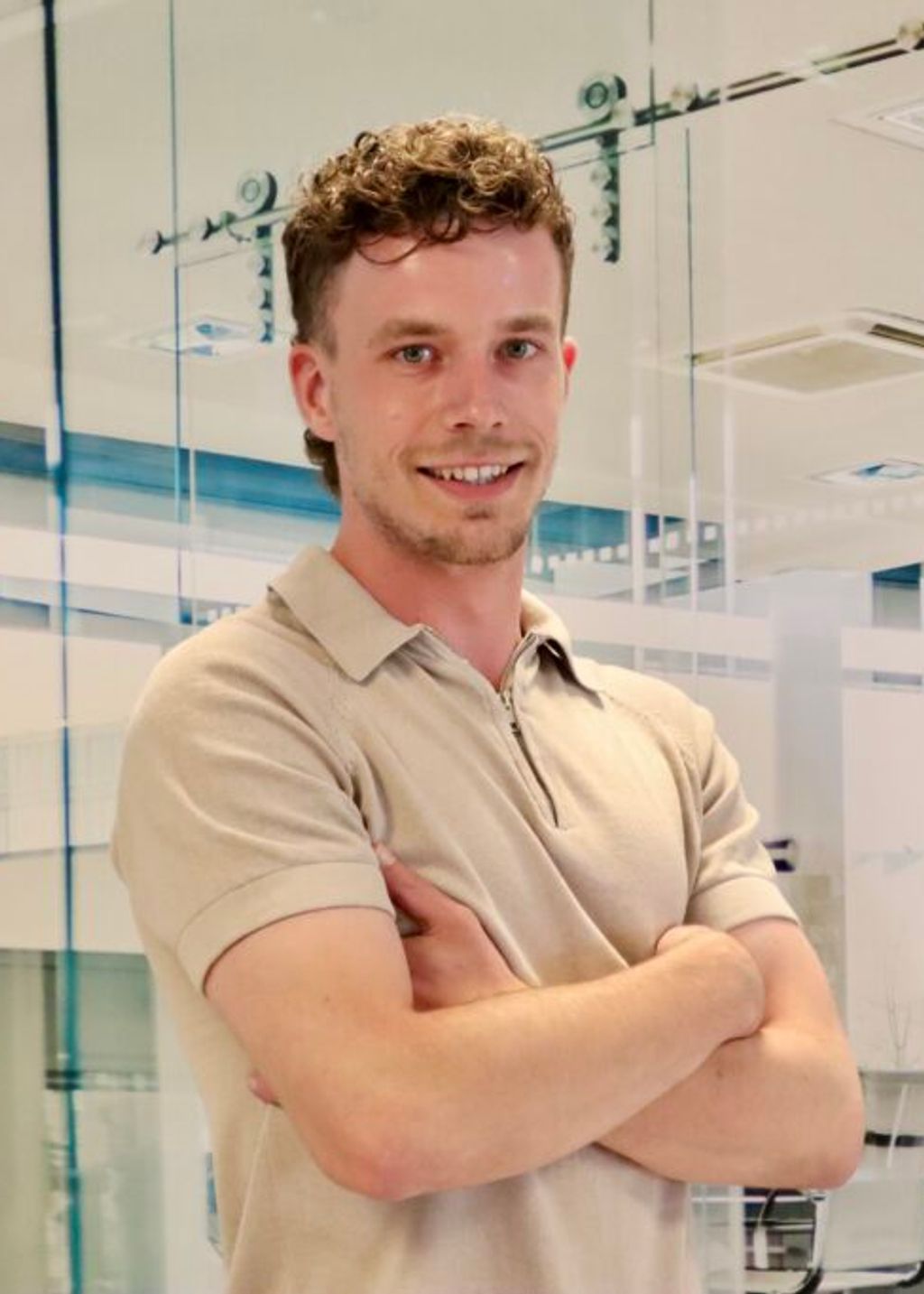 Man met donkerblond, krullend haar kijkt lachend de camera in met zijn armen over elkaar heen geslagen. Hij draagt een beige polo met korte mouwen.