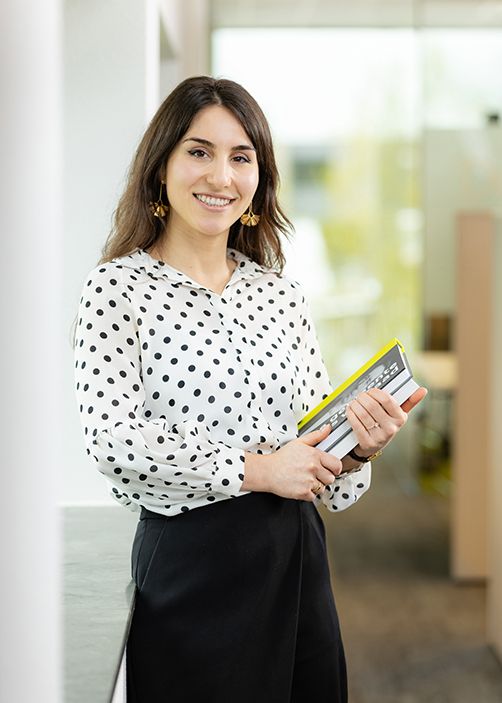 Portret van een jonge vrouw met een vriendelijke glimlach. Ze draagt een witte blouse met zwarte stippen en houdt een boek vast. Op de achtergrond is een moderne kantoorruimte te zien met veel daglicht.