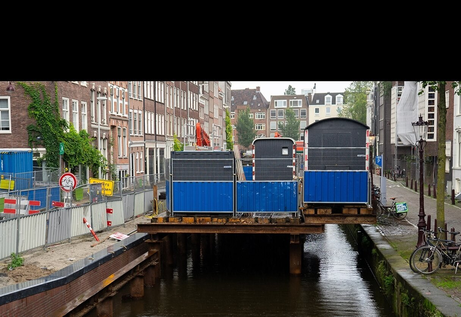 Renovatiewerkzaamheden aan een Amsterdamse kade met hekken, containers en bouwmaterialen boven het water in een smalle gracht in de binnenstad.