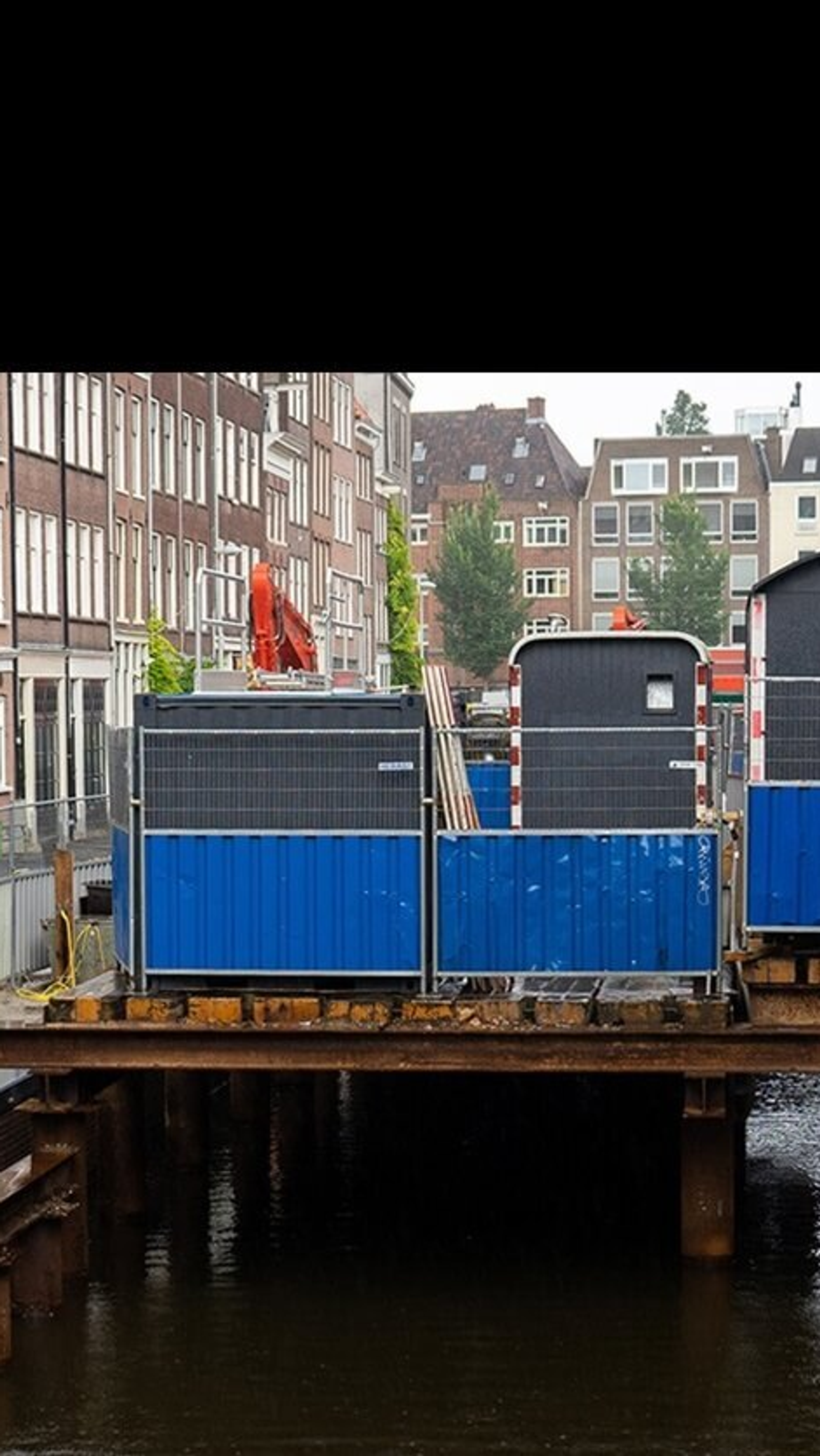 Renovatiewerkzaamheden aan een Amsterdamse kade met hekken, containers en bouwmaterialen boven het water in een smalle gracht in de binnenstad.