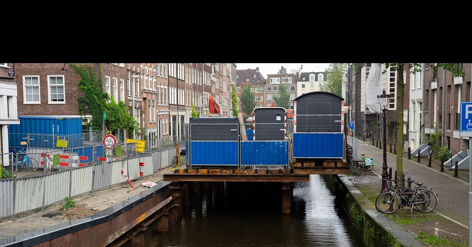 Renovatiewerkzaamheden aan een Amsterdamse kade met hekken, containers en bouwmaterialen boven het water in een smalle gracht in de binnenstad.