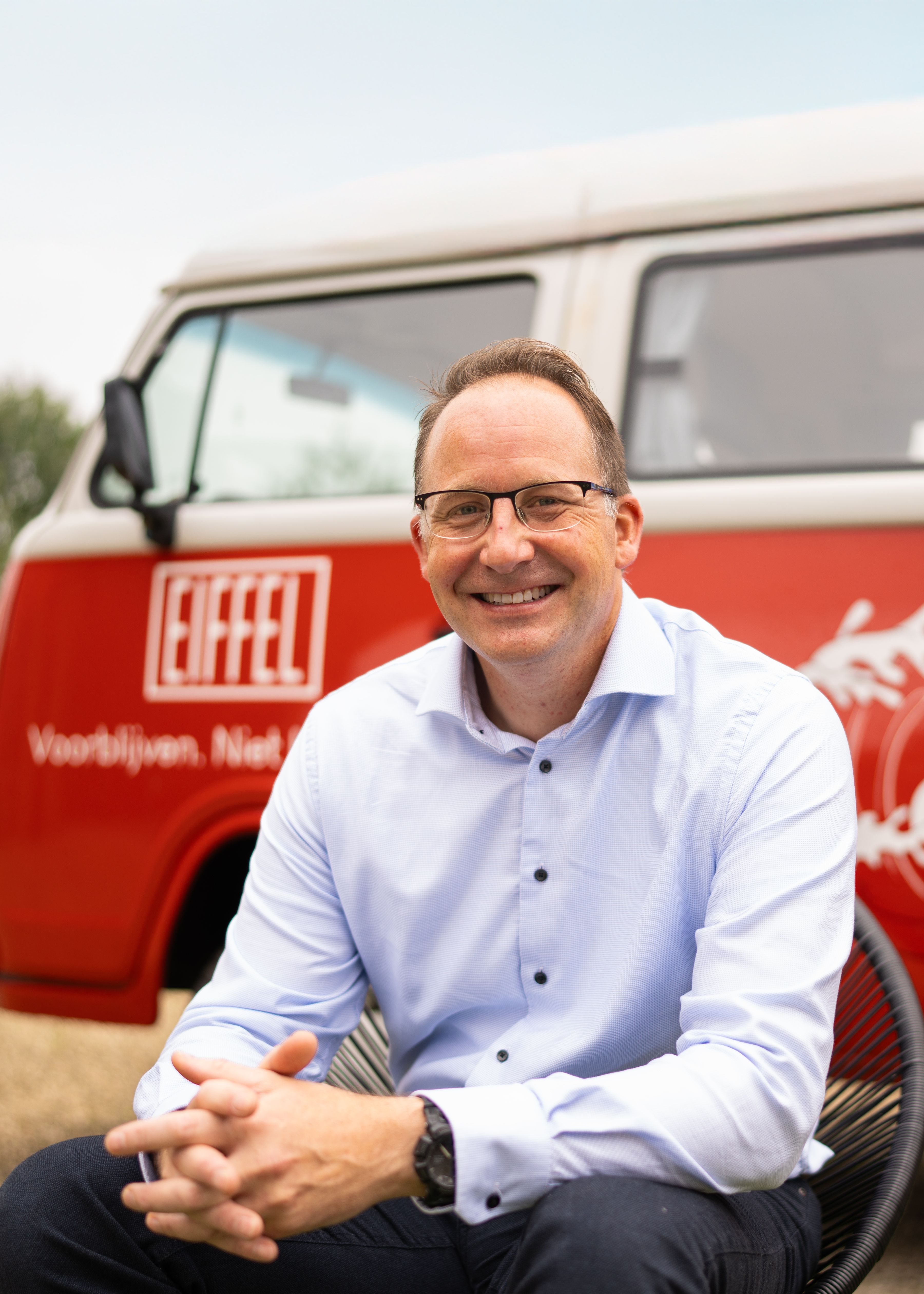 Een glimlachende man in een lichtblauw overhemd zit buiten op een stoel met een ontspannen houding. Op de achtergrond is een rode Volkswagen-bus te zien met het logo van EIFFEL.
