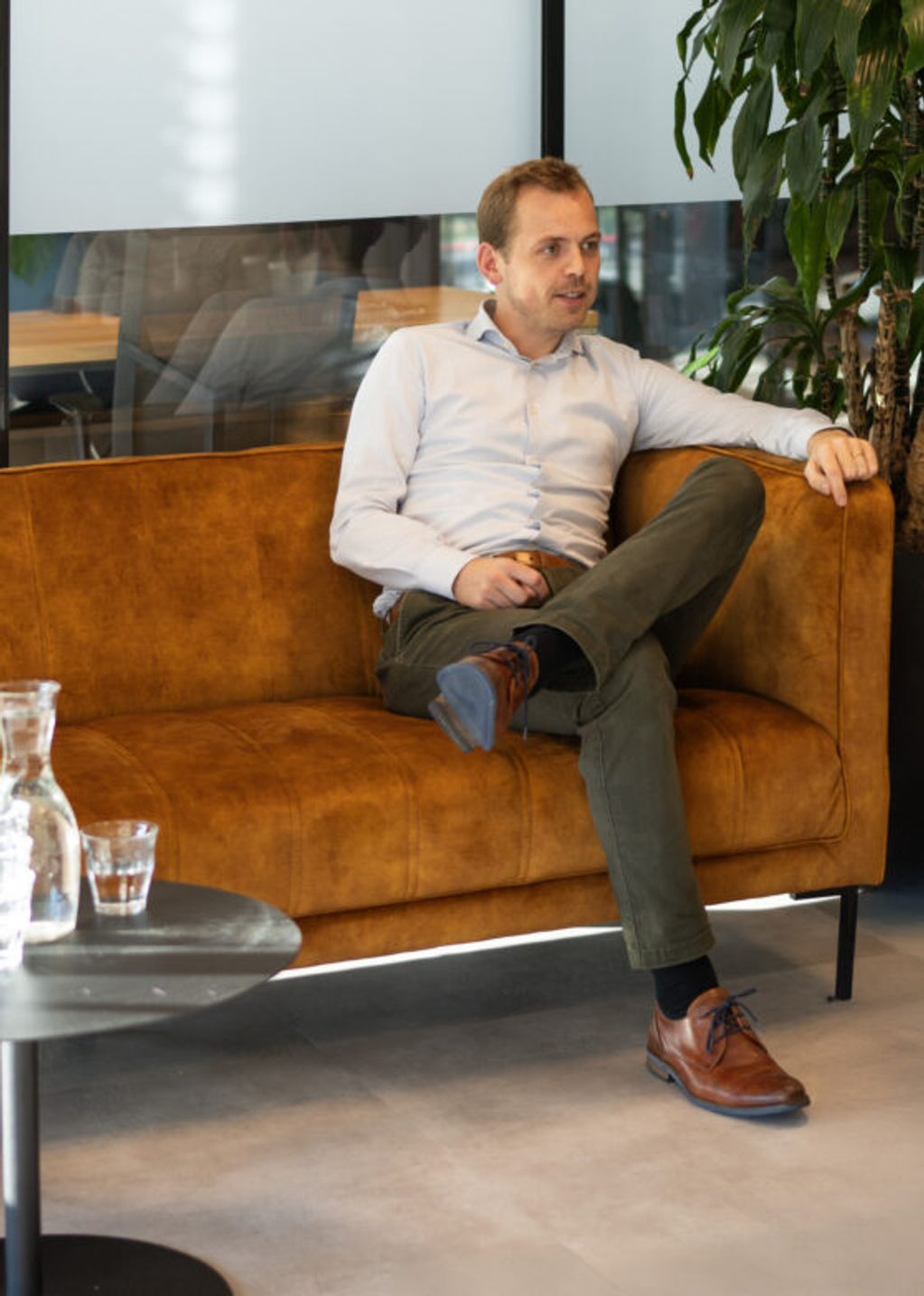 Twee mannen zitten op de bank terwijl ze in gesprek zijn met iemand buiten de camera. Eén van de mannen heeft bruin haar en draagt een beige broek met donkerblauwe polo. De andere man heeft donkerblond haar, draagt een donkere broek met licht overhemd.