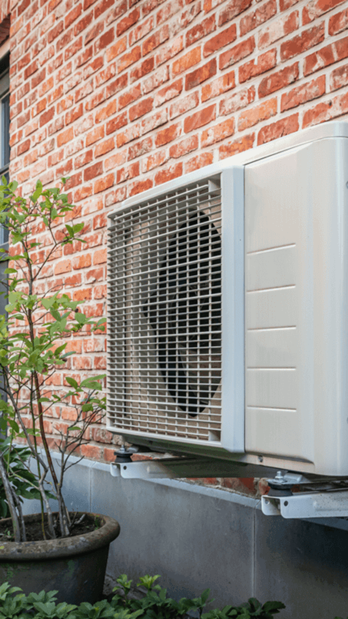 Warmtepompunit aan de buitenmuur van een huis met rode bakstenen, omgeven door groene planten en een tuinpad.