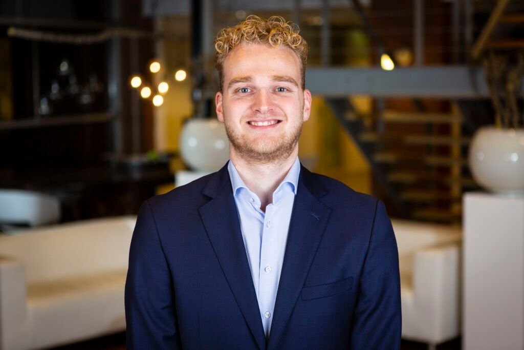 De man kijkt lachend de camera in, hij heeft blond krullen haar, blauwe ogen en een klein baardje. Hij draagt een lichtblauw overhemd met daarover een blauw jasje.