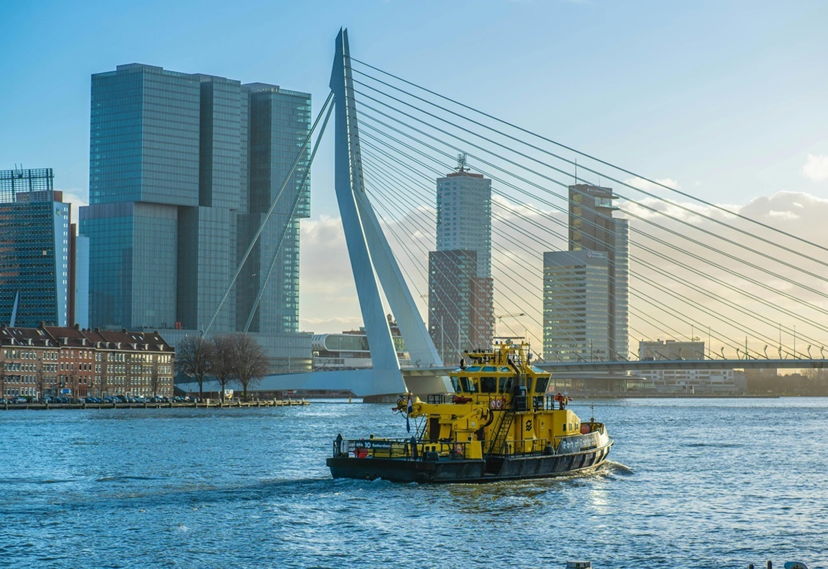 Gele havenboot vaart over de Maas in Rotterdam met op de achtergrond de Erasmusbrug en moderne hoogbouw aan de Kop van Zuid.
