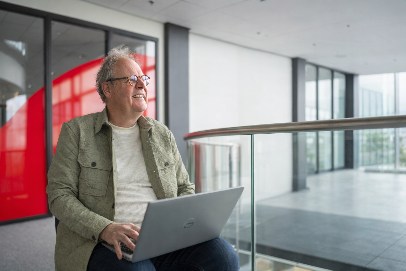 Een meneer met grijs haar zit op een poef en heeft zijn laptop op schoot, hij lacht naar iemand voorbij de camera