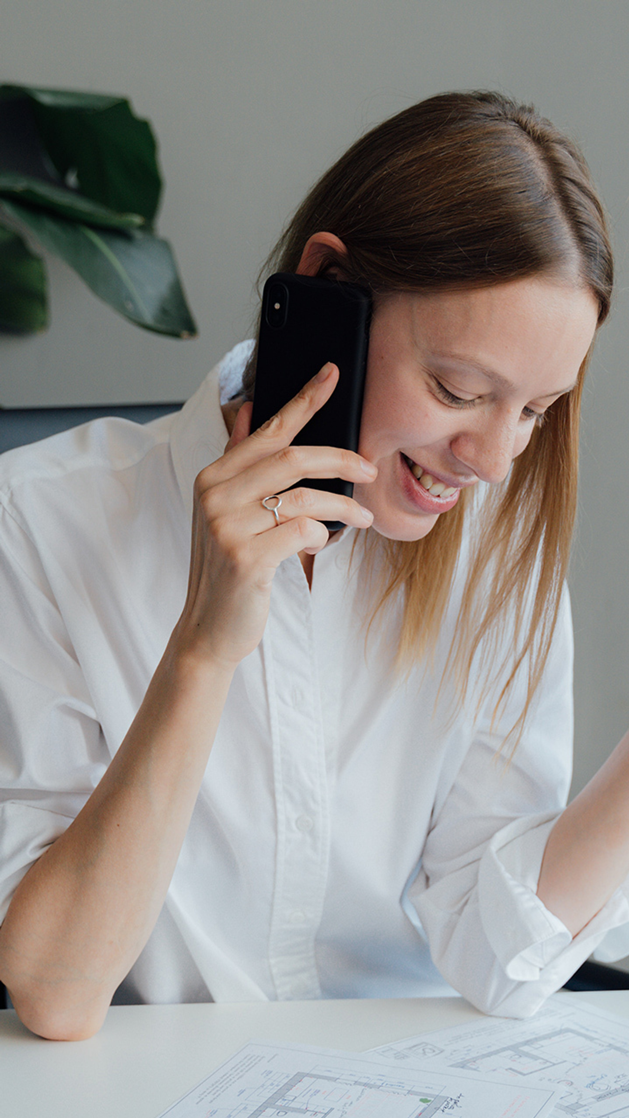 Vrouw in witte blouse voert een telefoongesprek terwijl ze lacht, met bouwtekeningen voor zich op tafel en een pen in haar hand.