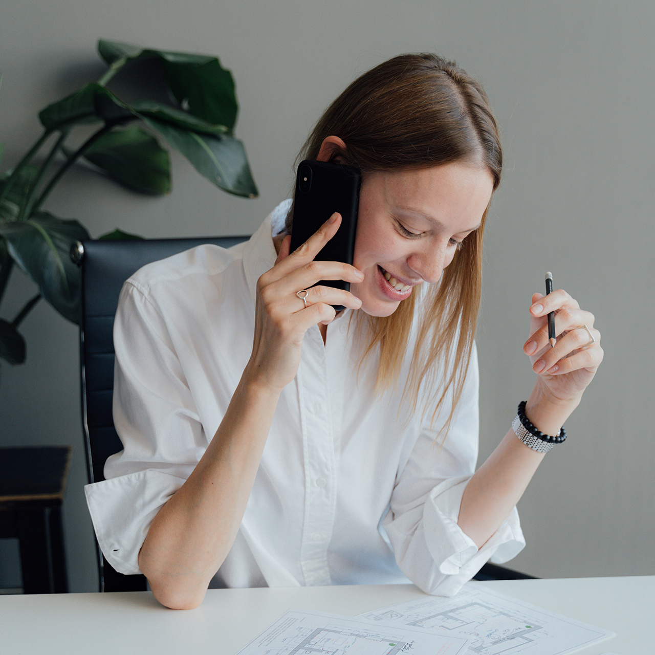 Vrouw in witte blouse voert een telefoongesprek terwijl ze lacht, met bouwtekeningen voor zich op tafel en een pen in haar hand.