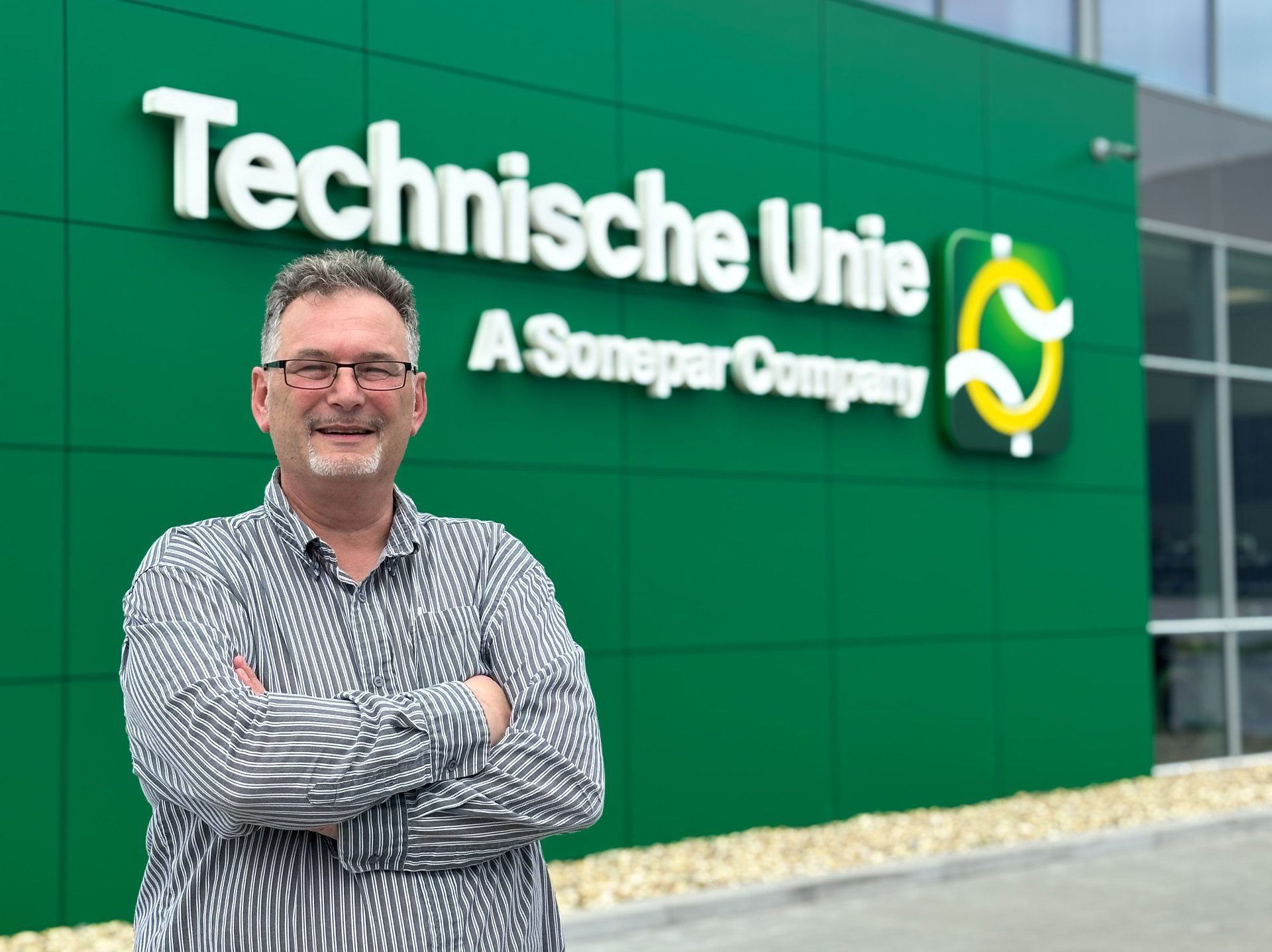 Man met blouse, armen over elkaar, lachend voor het gebouw van de Technische Unie