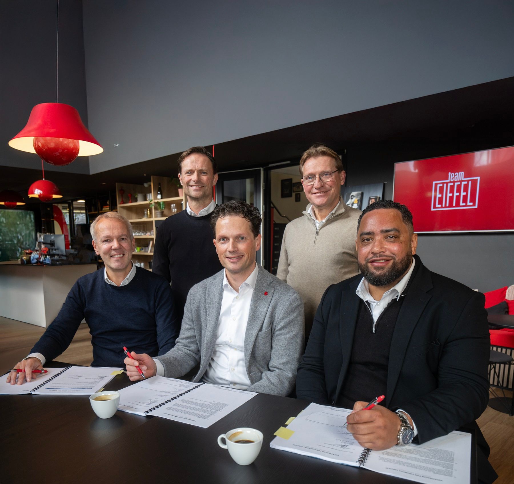 Vijf mannen poseren lachend rondom een tafel bij Team EIFFEL. Drie van hen zitten aan tafel met open documenten en rode pennen in de hand, alsof ze zojuist iets ondertekend hebben. Twee mannen staan achter hen. Op de achtergrond zijn een koffietafel, boekenkast en het Team EIFFEL-logo op een scherm zichtbaar.