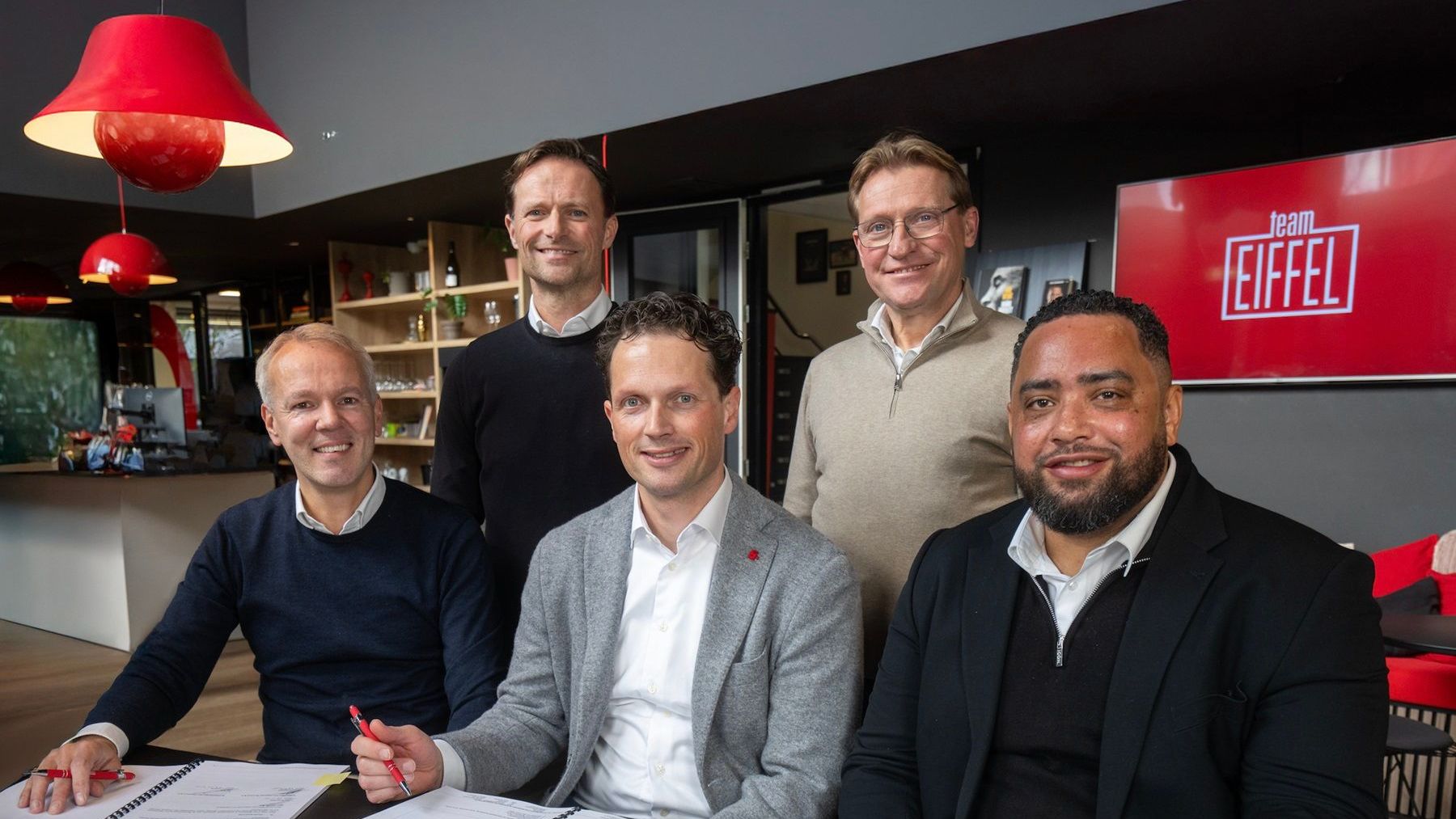 Vijf mannen poseren lachend rondom een tafel bij Team EIFFEL. Drie van hen zitten aan tafel met open documenten en rode pennen in de hand, alsof ze zojuist iets ondertekend hebben. Twee mannen staan achter hen. Op de achtergrond zijn een koffietafel, boekenkast en het Team EIFFEL-logo op een scherm zichtbaar.