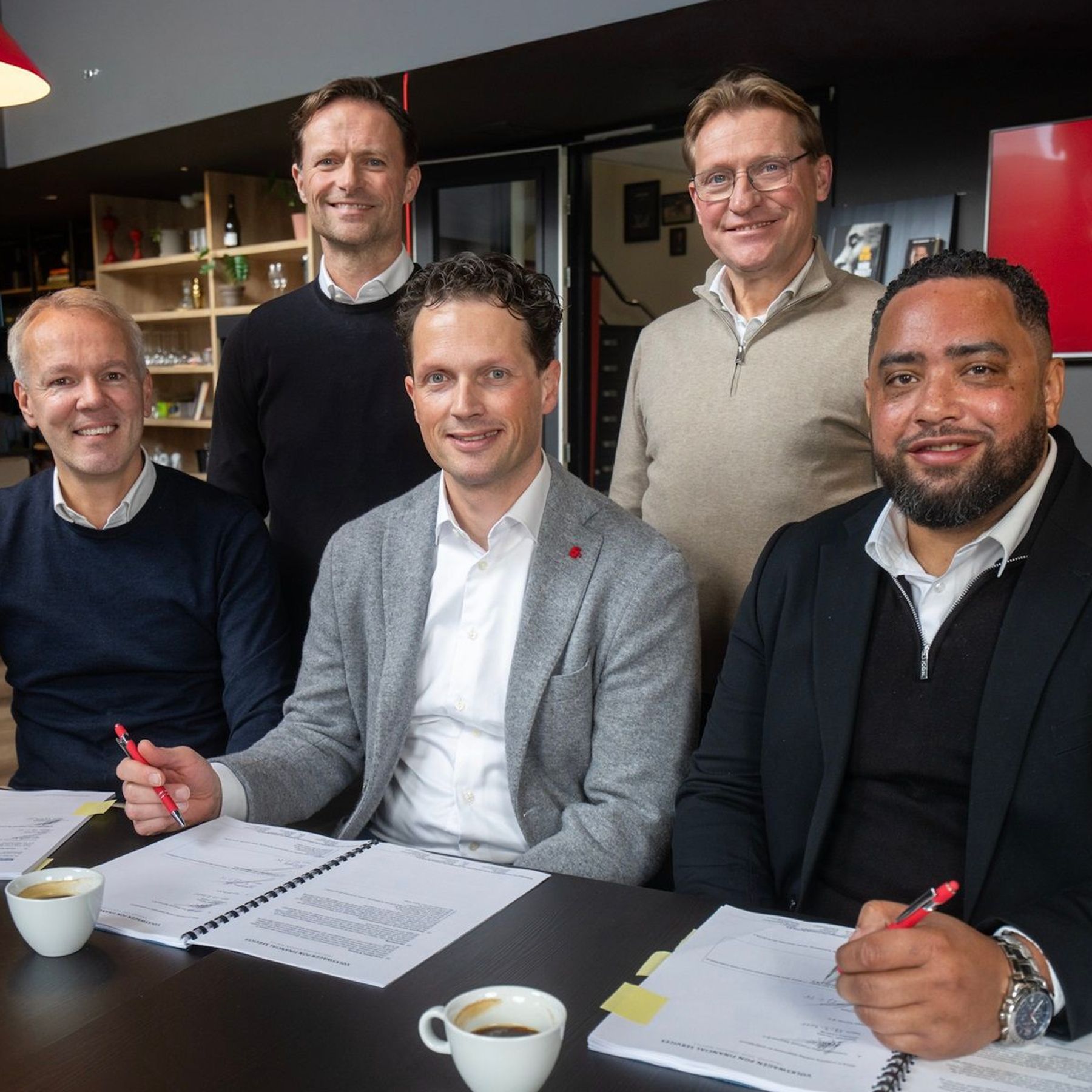 Vijf mannen poseren lachend rondom een tafel bij Team EIFFEL. Drie van hen zitten aan tafel met open documenten en rode pennen in de hand, alsof ze zojuist iets ondertekend hebben. Twee mannen staan achter hen. Op de achtergrond zijn een koffietafel, boekenkast en het Team EIFFEL-logo op een scherm zichtbaar.