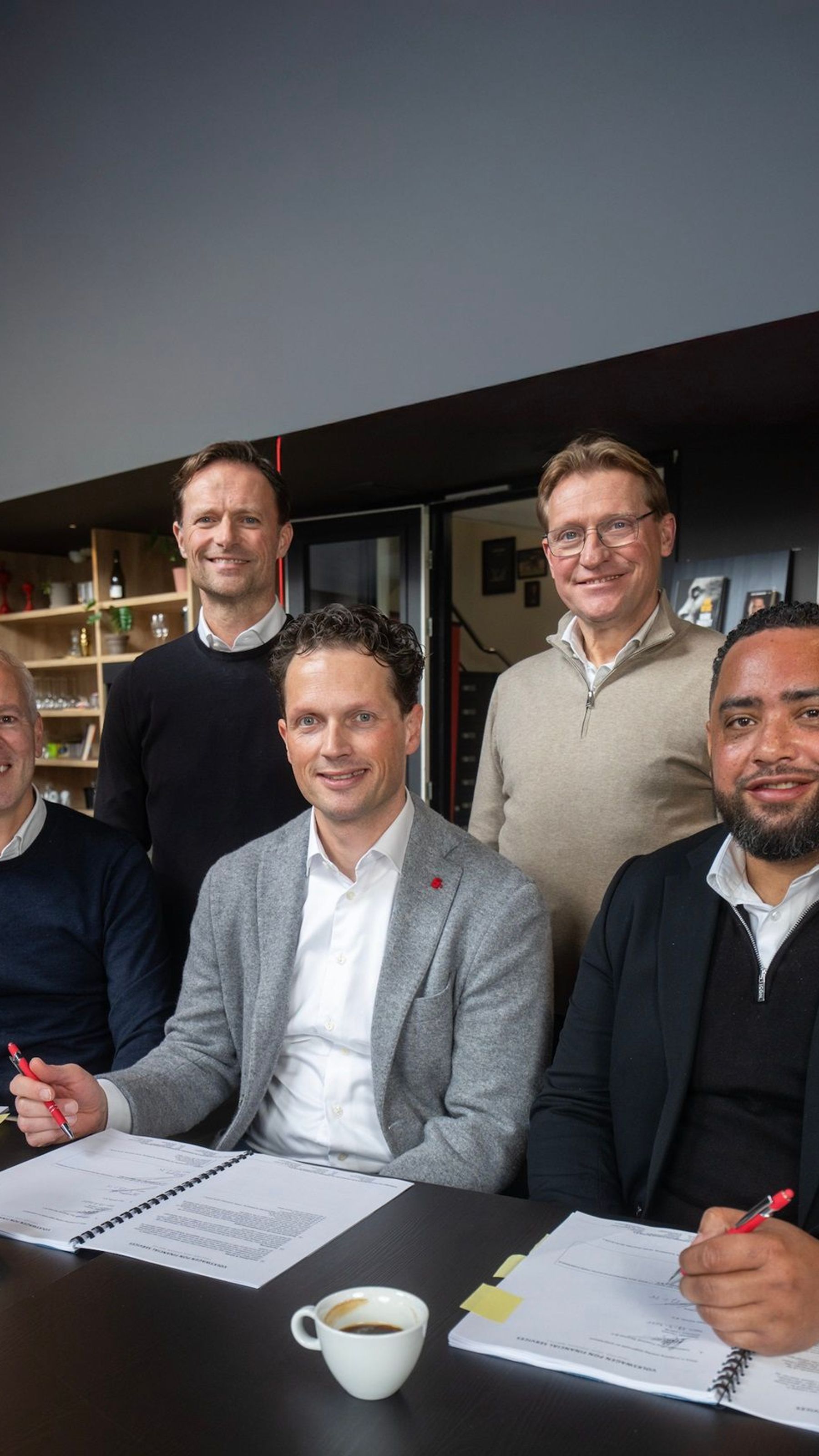 Vijf mannen poseren lachend rondom een tafel bij Team EIFFEL. Drie van hen zitten aan tafel met open documenten en rode pennen in de hand, alsof ze zojuist iets ondertekend hebben. Twee mannen staan achter hen. Op de achtergrond zijn een koffietafel, boekenkast en het Team EIFFEL-logo op een scherm zichtbaar.