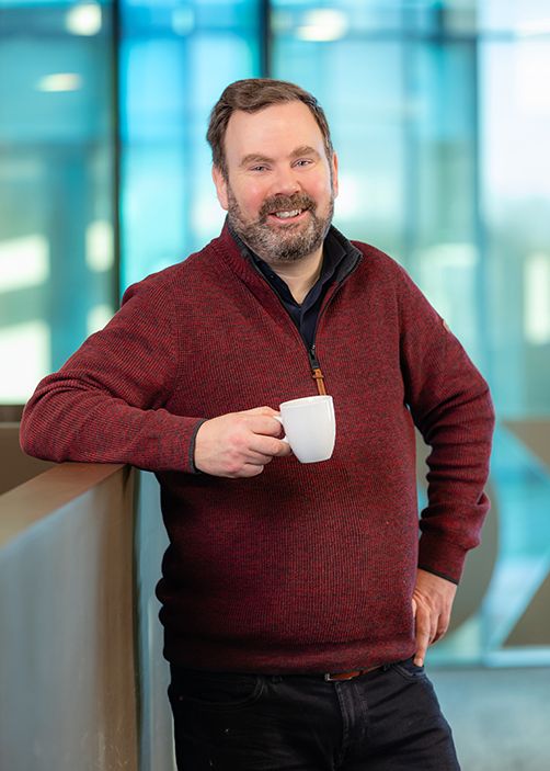 Portret van een man met een vriendelijke glimlach, leunend tegen een balustrade en een witte mok in zijn hand. Hij draagt een donkerrode trui en poseert in een moderne, lichtovergoten binnenruimte met glazen wanden.