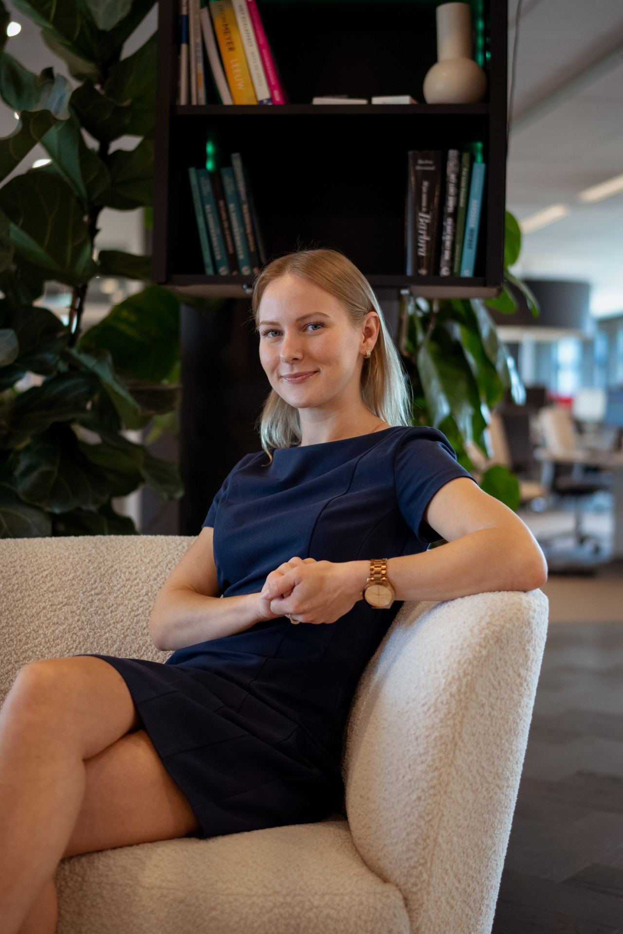Blonde vrouw, zittend op een stoffen bankje. Draagt een blauwe nette jurk tot boven de knie en lacht vriendelijk naar de camera.