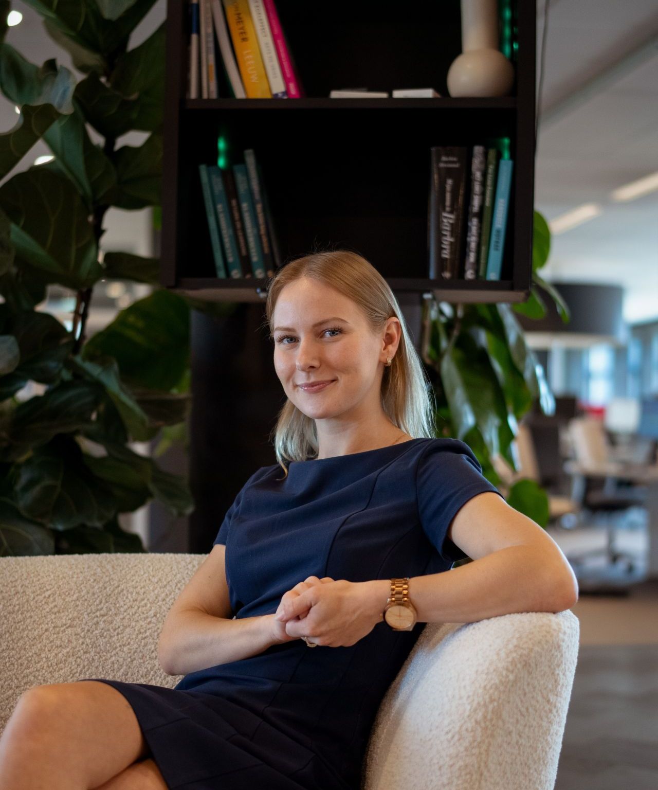 Blonde vrouw, zittend op een stoffen bankje. Draagt een blauwe nette jurk tot boven de knie en lacht vriendelijk naar de camera.