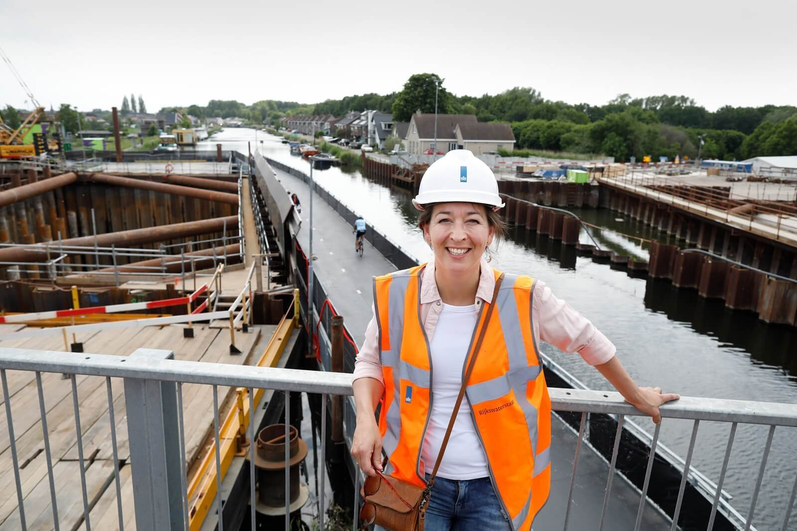 Omgevingsadviseur Nienke in veiligheidshesje en helm op een bouwlocatie van de A16 Rotterdam, met zicht op werkzaamheden langs het water.