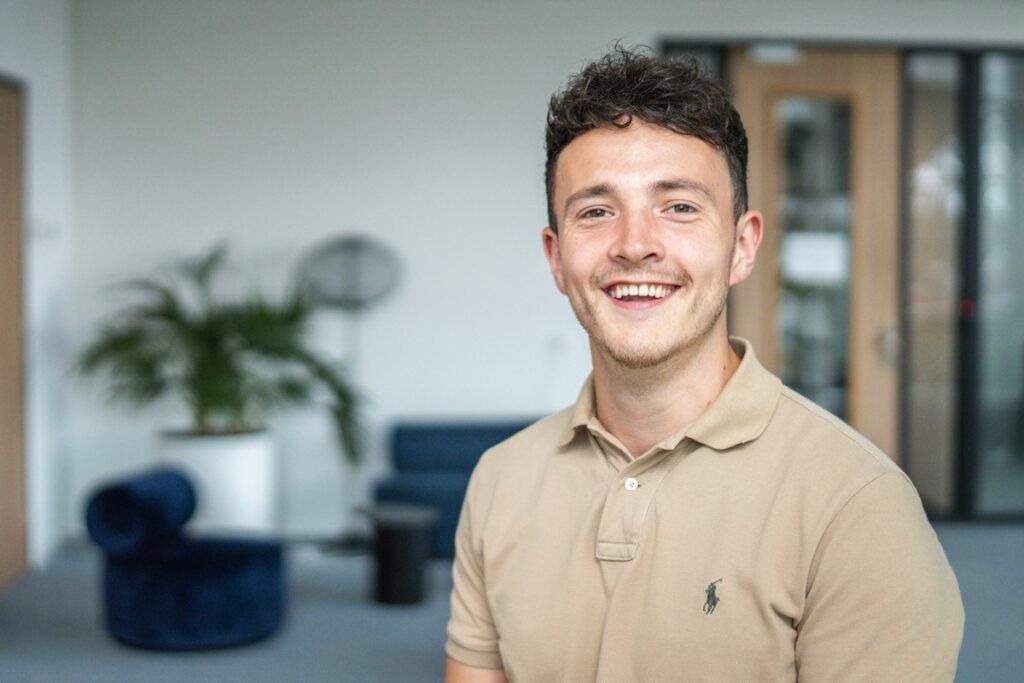 Man kijkt lachend in de camera, heeft bruin krullend haar en draagt een beige polo