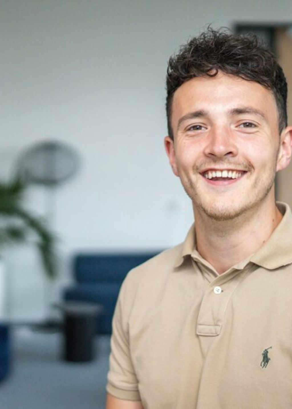 Man kijkt lachend in de camera, heeft bruin krullend haar en draagt een beige polo