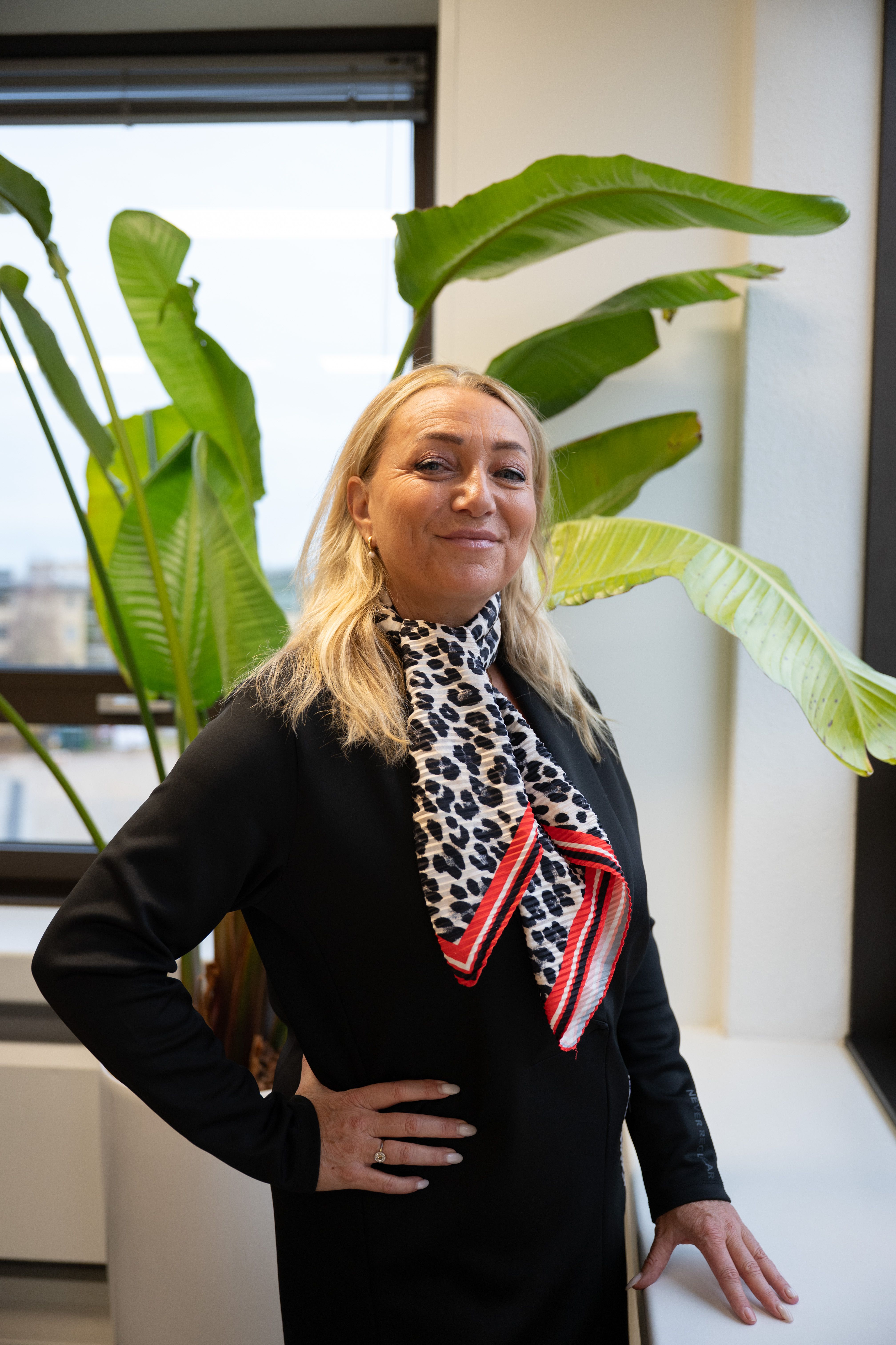 Blonde vrouw lacht naar de camera, ze draagt een zwarte colbert en een panterprint sjaaltje.