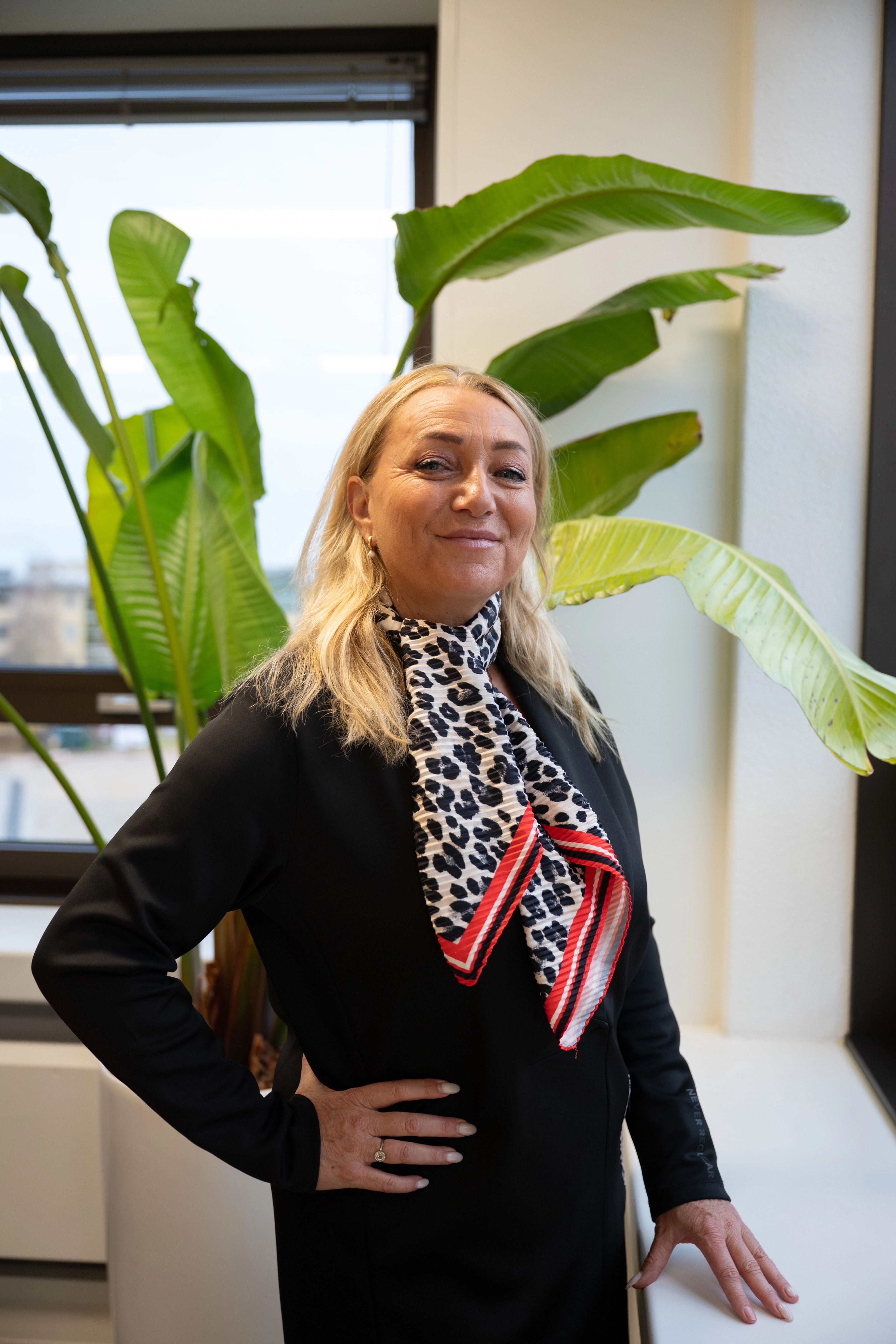 Blonde vrouw lacht naar de camera, ze draagt een zwarte colbert en een panterprint sjaaltje.