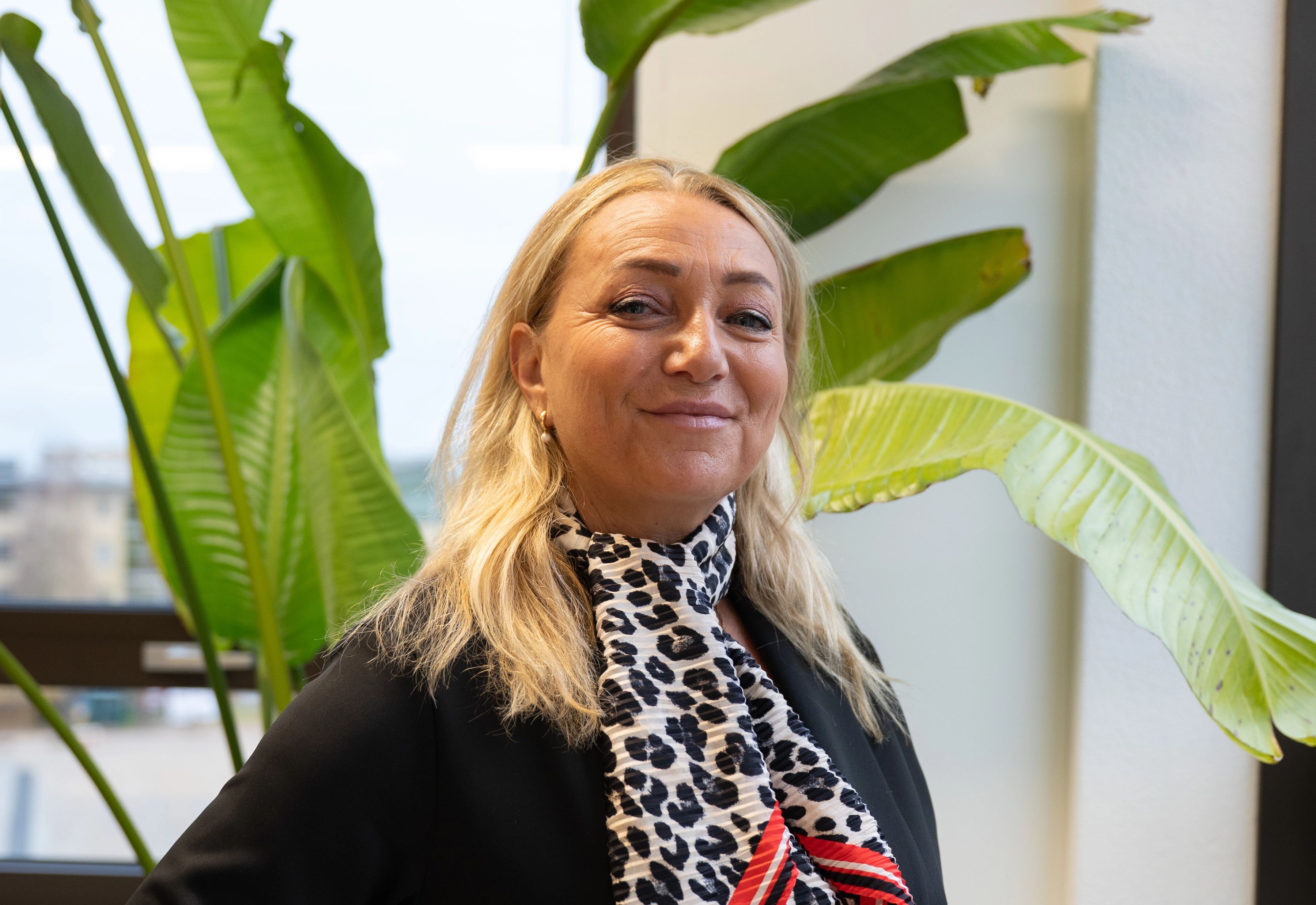 Blonde vrouw lacht naar de camera, ze draagt een zwarte colbert en een panterprint sjaaltje.