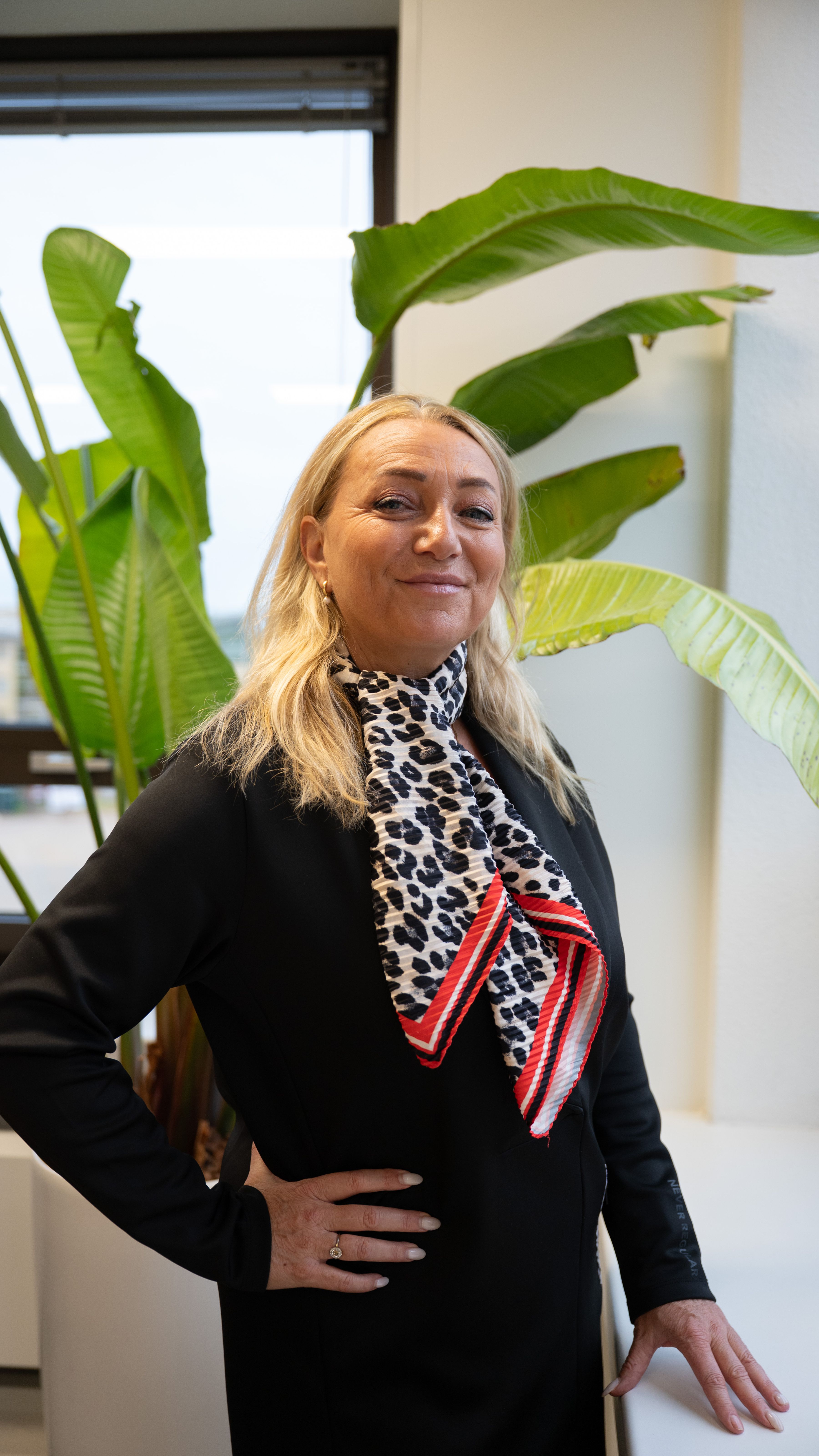 Blonde vrouw lacht naar de camera, ze draagt een zwarte colbert en een panterprint sjaaltje.