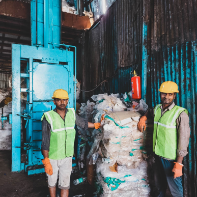Two men wearing safety vests and helmets standing next to a pile of garbage.