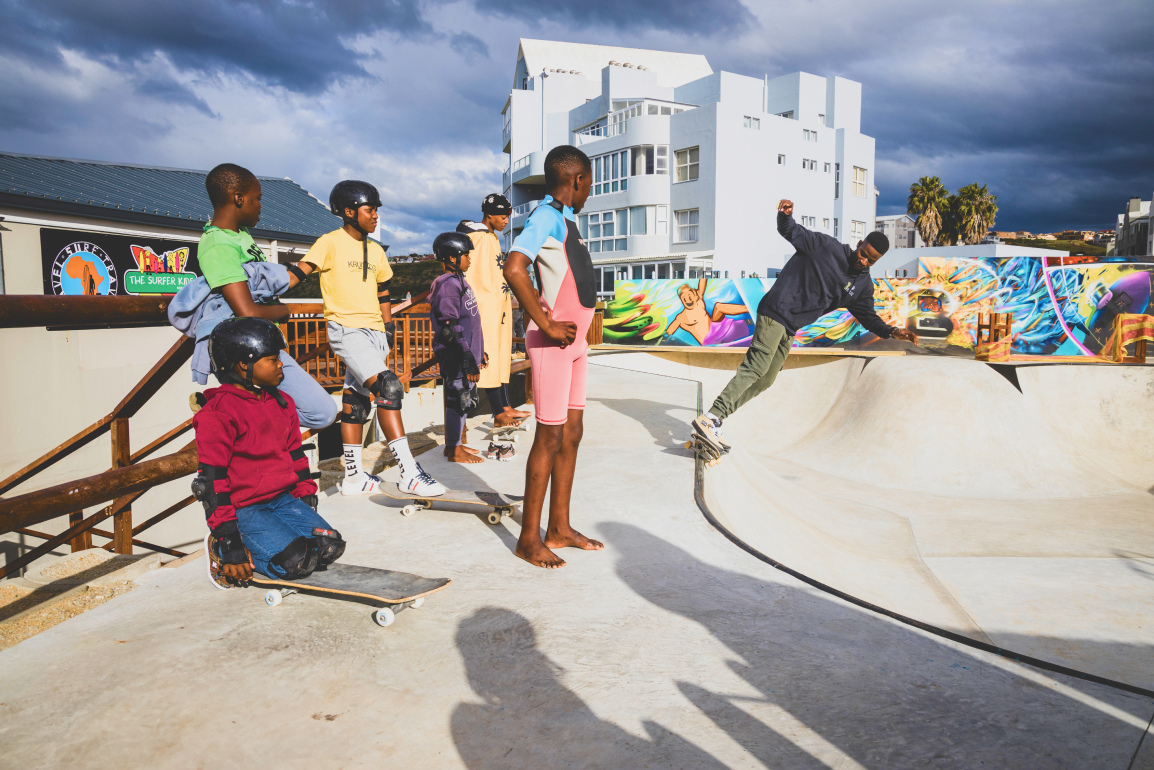 A group of children watch another child ride a skateboard in a skatepark.