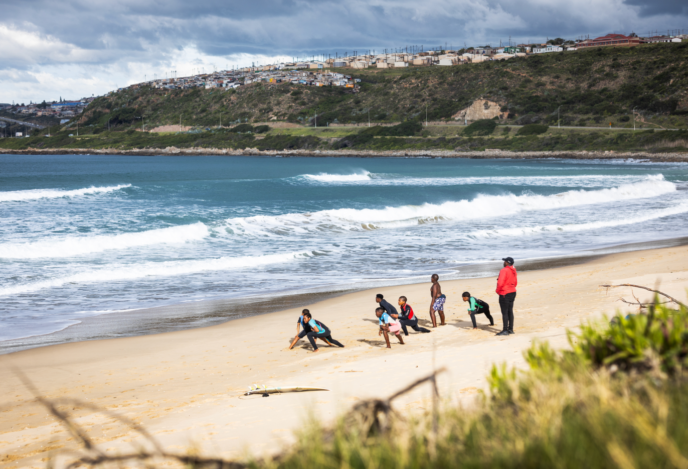 A group of children stands stretching their legs on a sand beach beside an ocean.