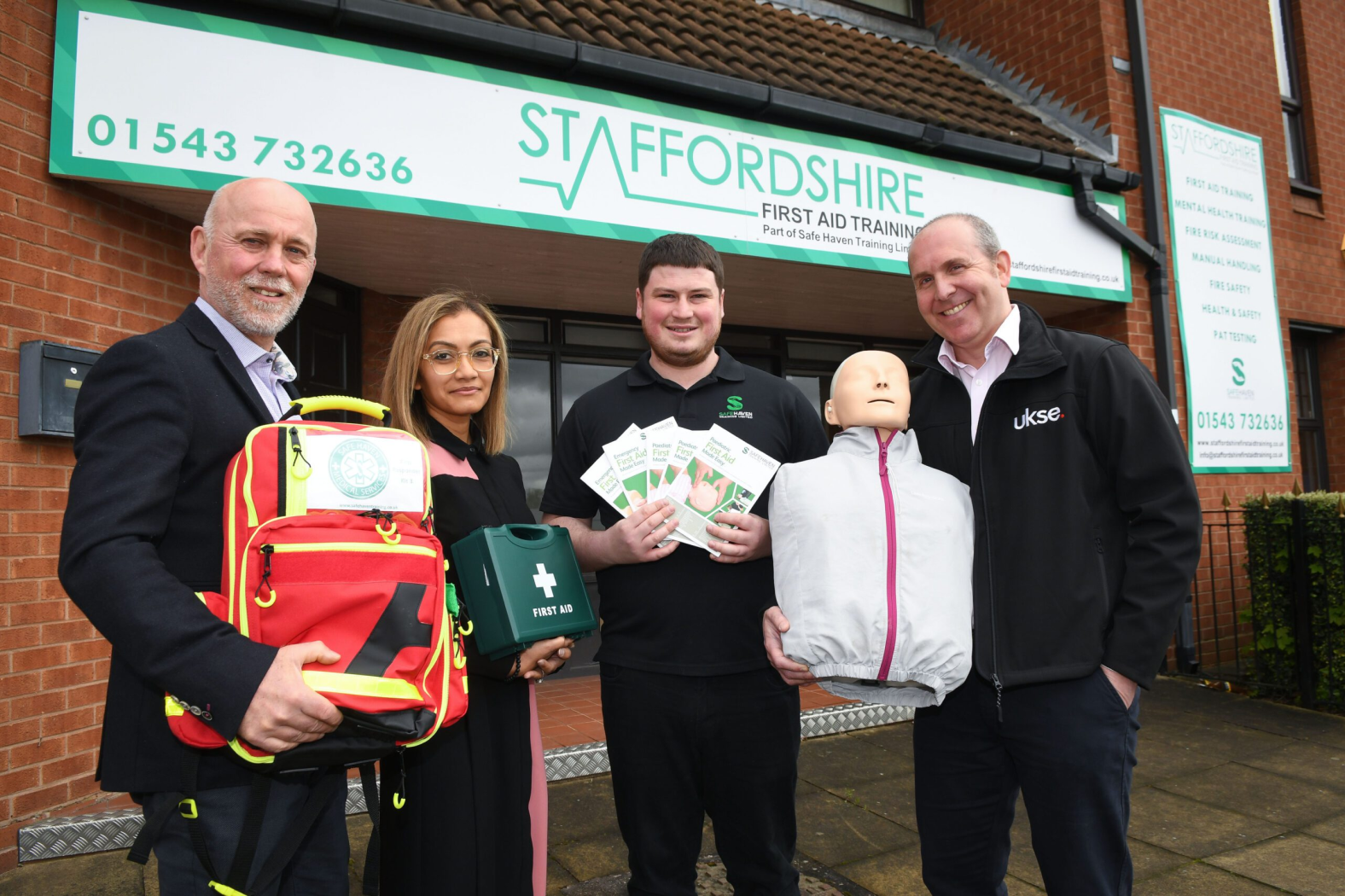 Four people holding first aid items stand outside a first aid training clinic.