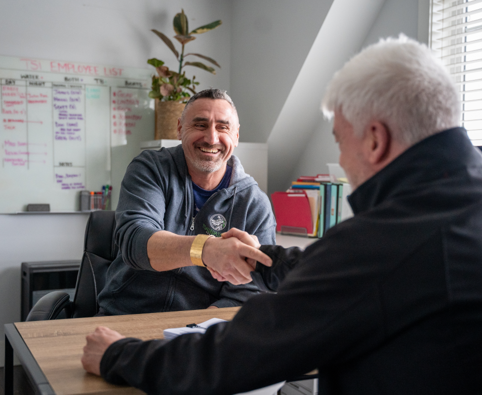 Two people shake hands sitting across from each other at a desk.