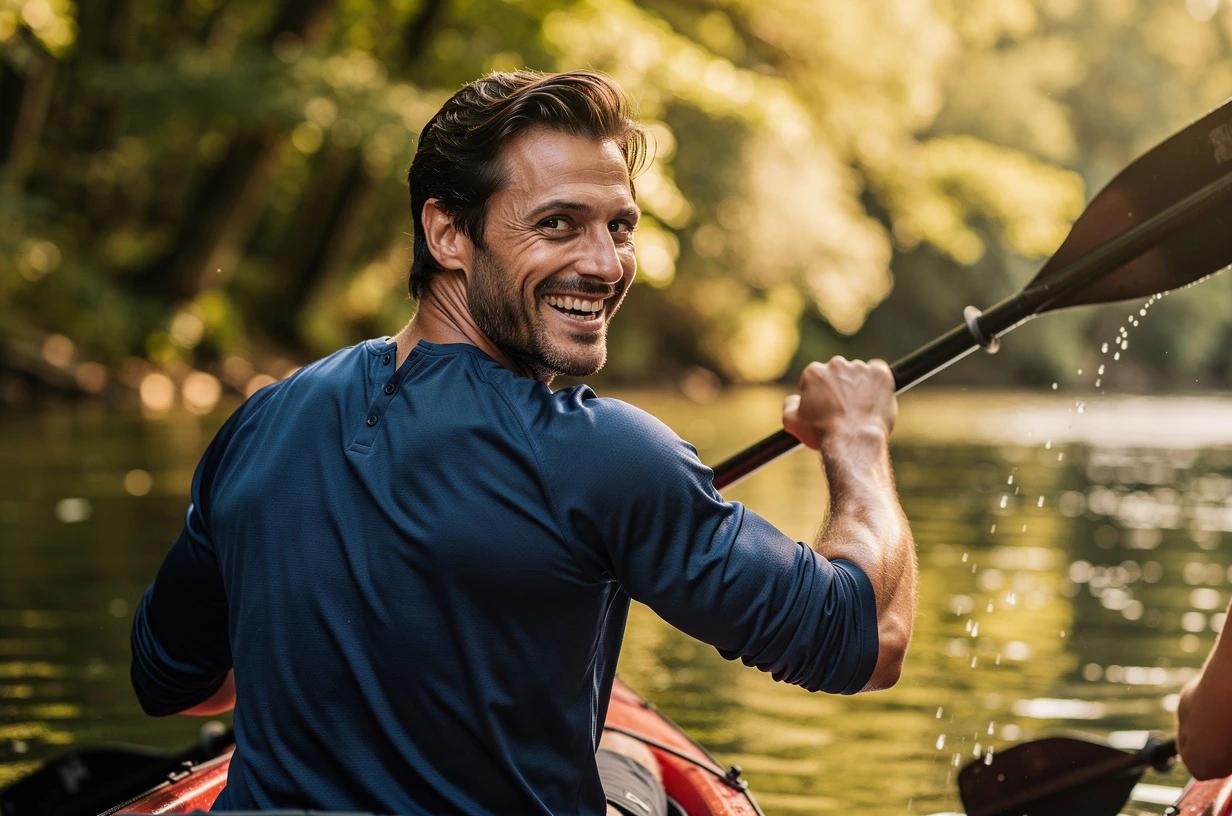 A man in his mid-30s paddles a kayak through calm river water, laughing and glancing back over his shoulder at a companion just out of frame.