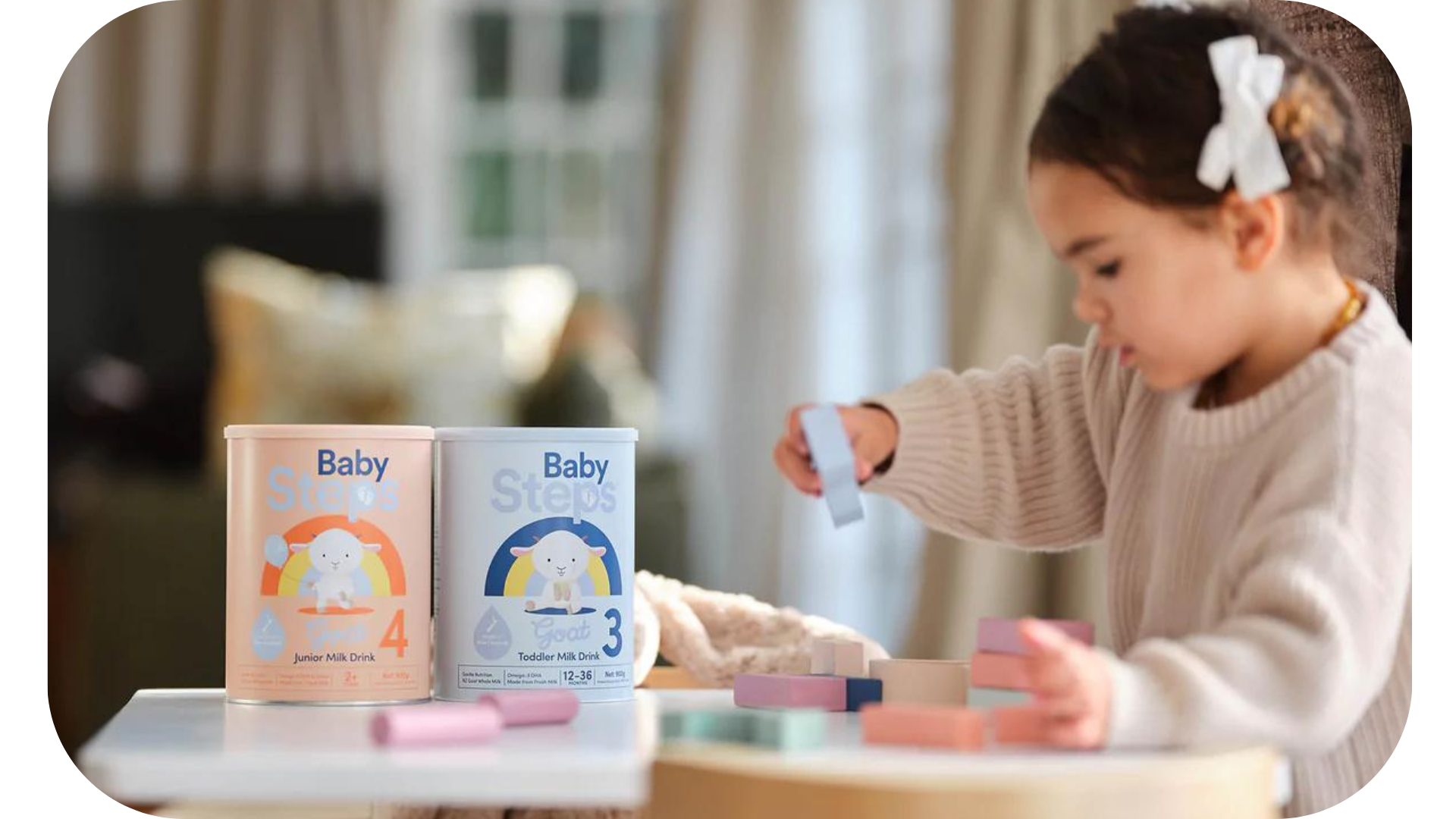 A young girl engaging in play with building blocks, with two cans of Baby Steps, a goat's milk drink for children, prominently displayed in the background.