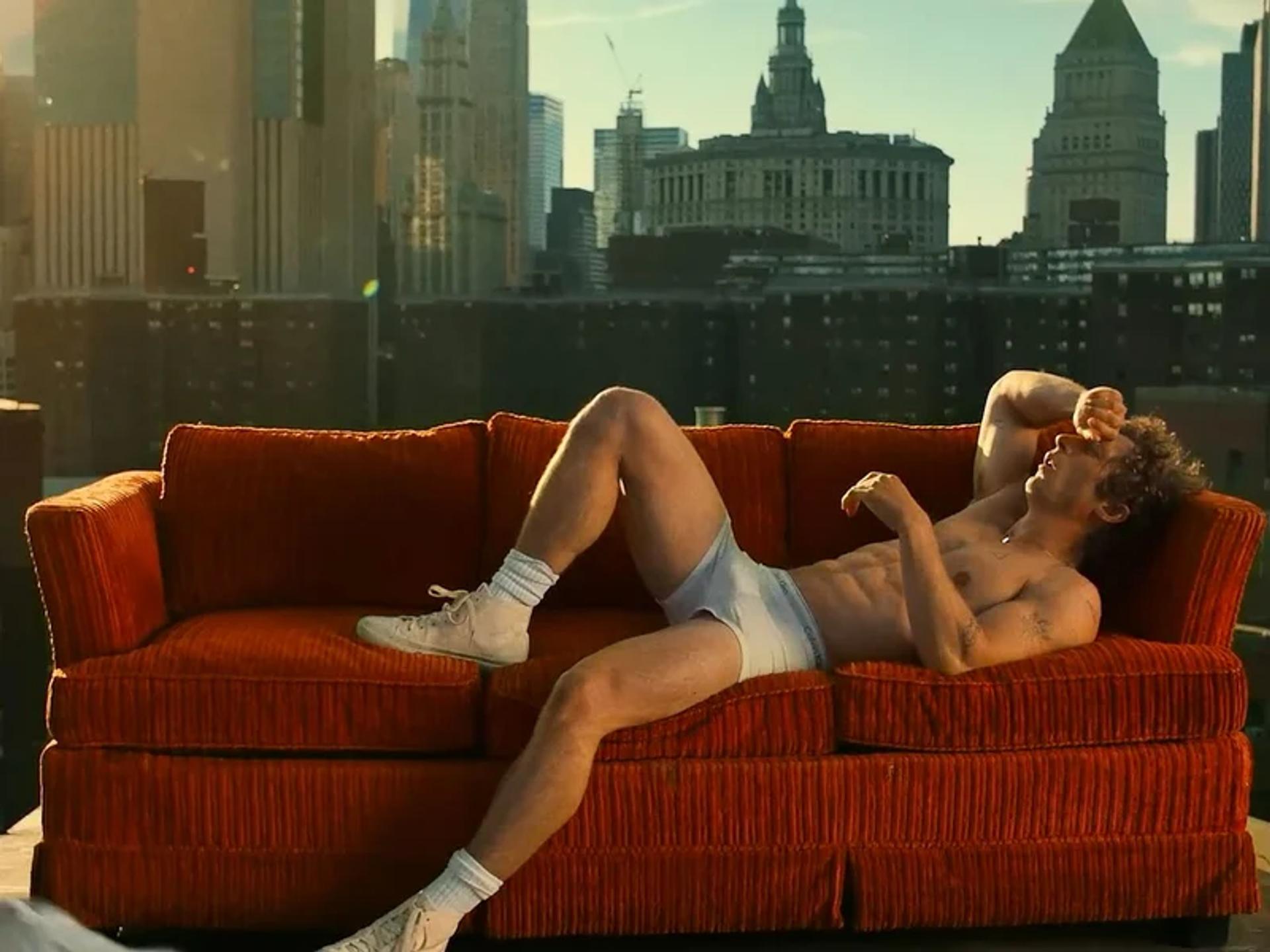 A shirtless man lounging on a red couch in a room with a city skyline view, representing the Jeremy White Allen's Calvin Klein campaign