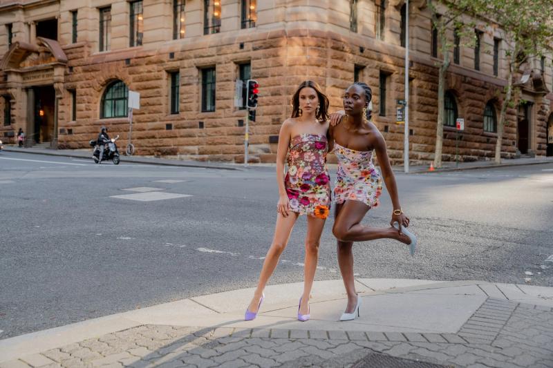 Two women in colorful strapless floral mini-dresses pose on a city street corner.