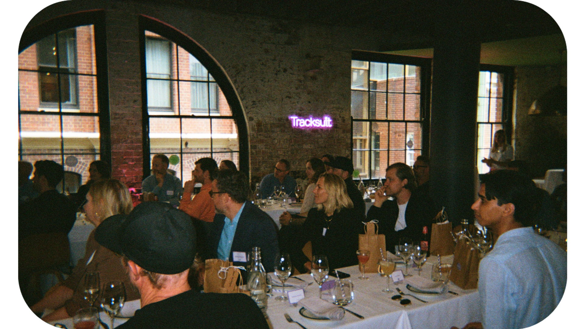 A group of people engaged in a discussion at tables, with a neon sign displaying the word 'Tracksuit' in the background. The image is from a marketing event where Mark Ritson shared insights on brand strategy and marketing.