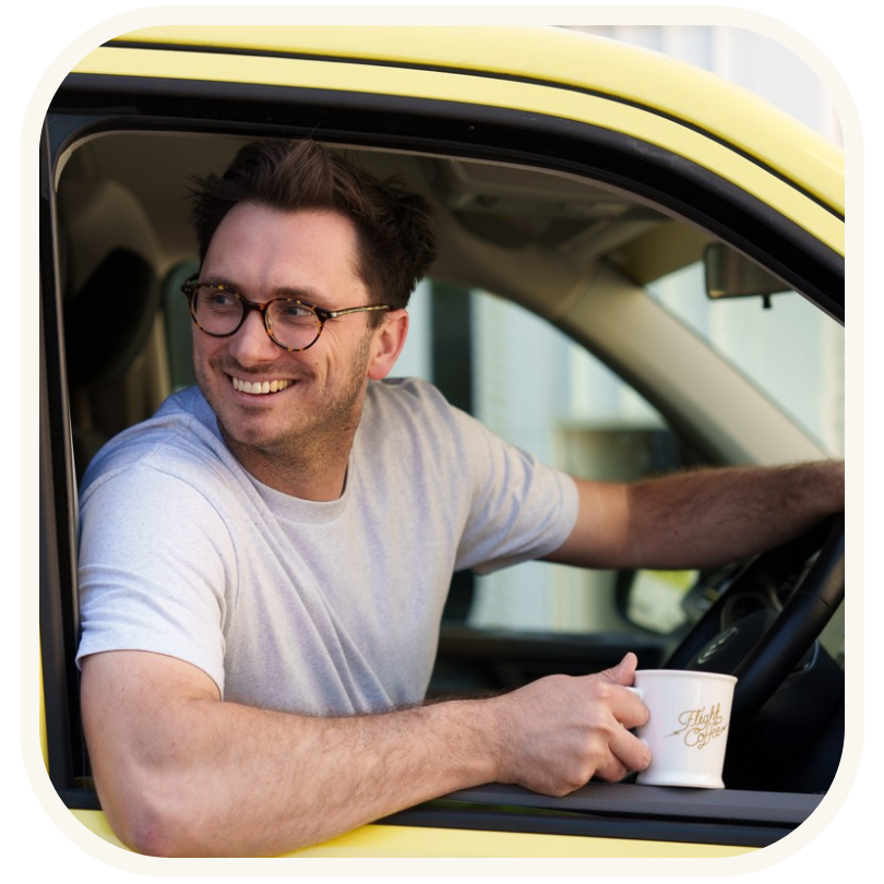 A man in a yellow van, representing Flight Coffee, holding a can of their Oat Milk Flat White coffee, ready to enjoy it. This image is part of a coffee advertising campaign.