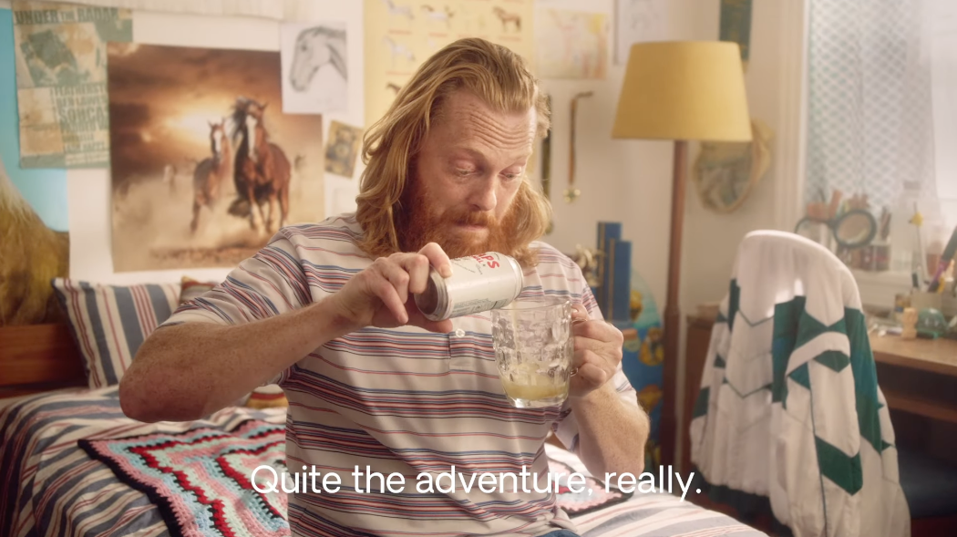 A man sitting on a bed, pouring a non-alcoholic Heaps Normal beer into a mug, with the caption 'quite the adventure really' below him, representing the brand's fun and adventurous spirit