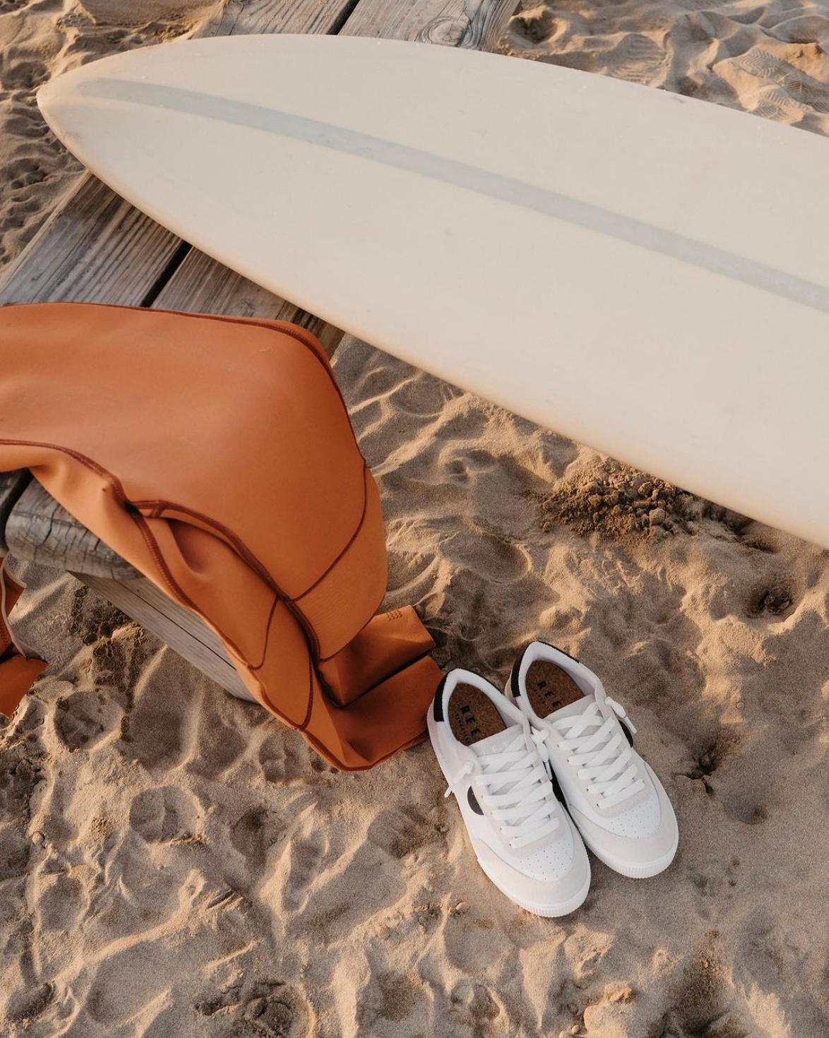 A pair of Reef brand shoes sitting on the sand next to a surfboard