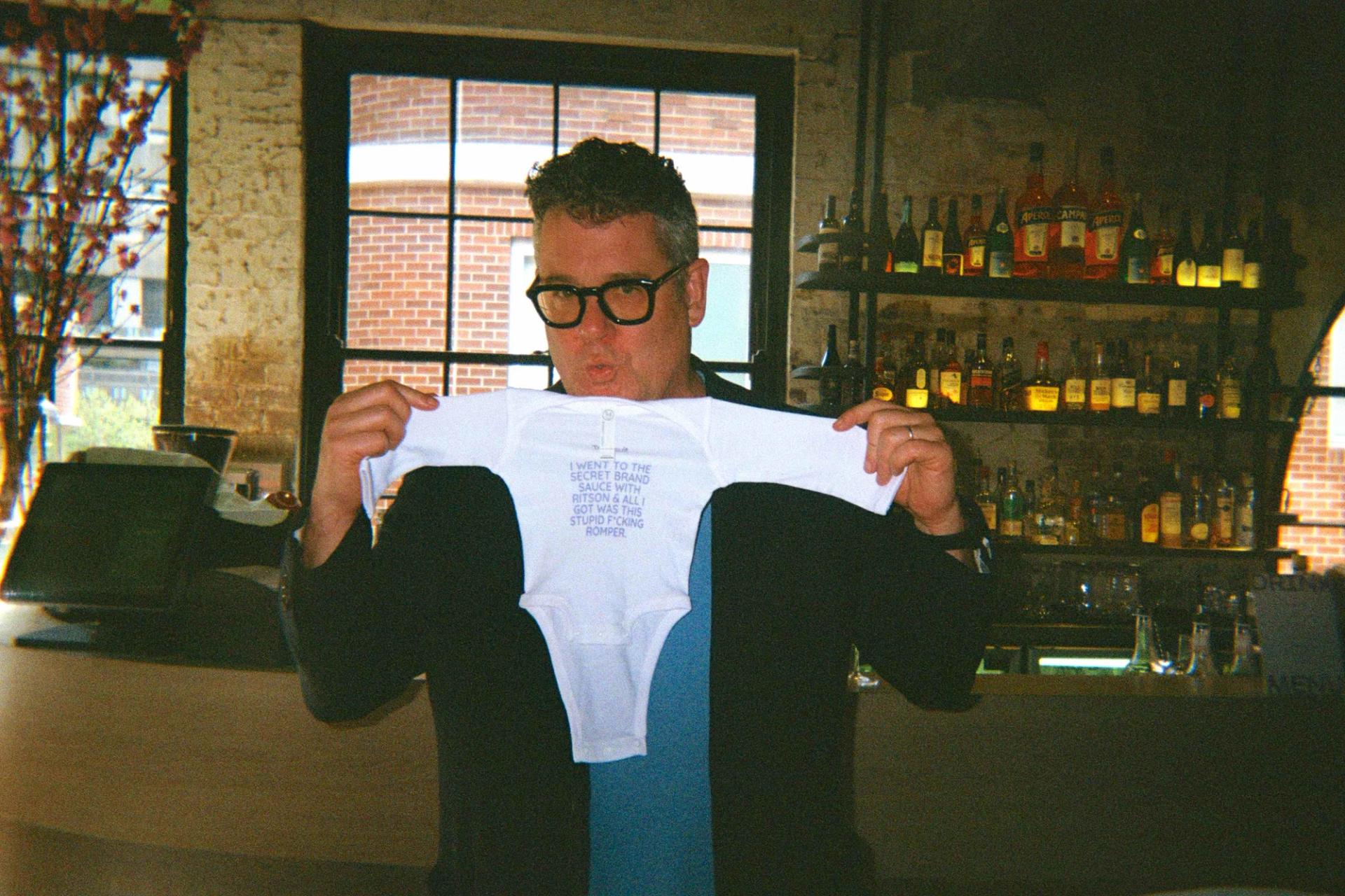 mark ritson holds up a Tracksuit branded baby playsuit in a classy bar