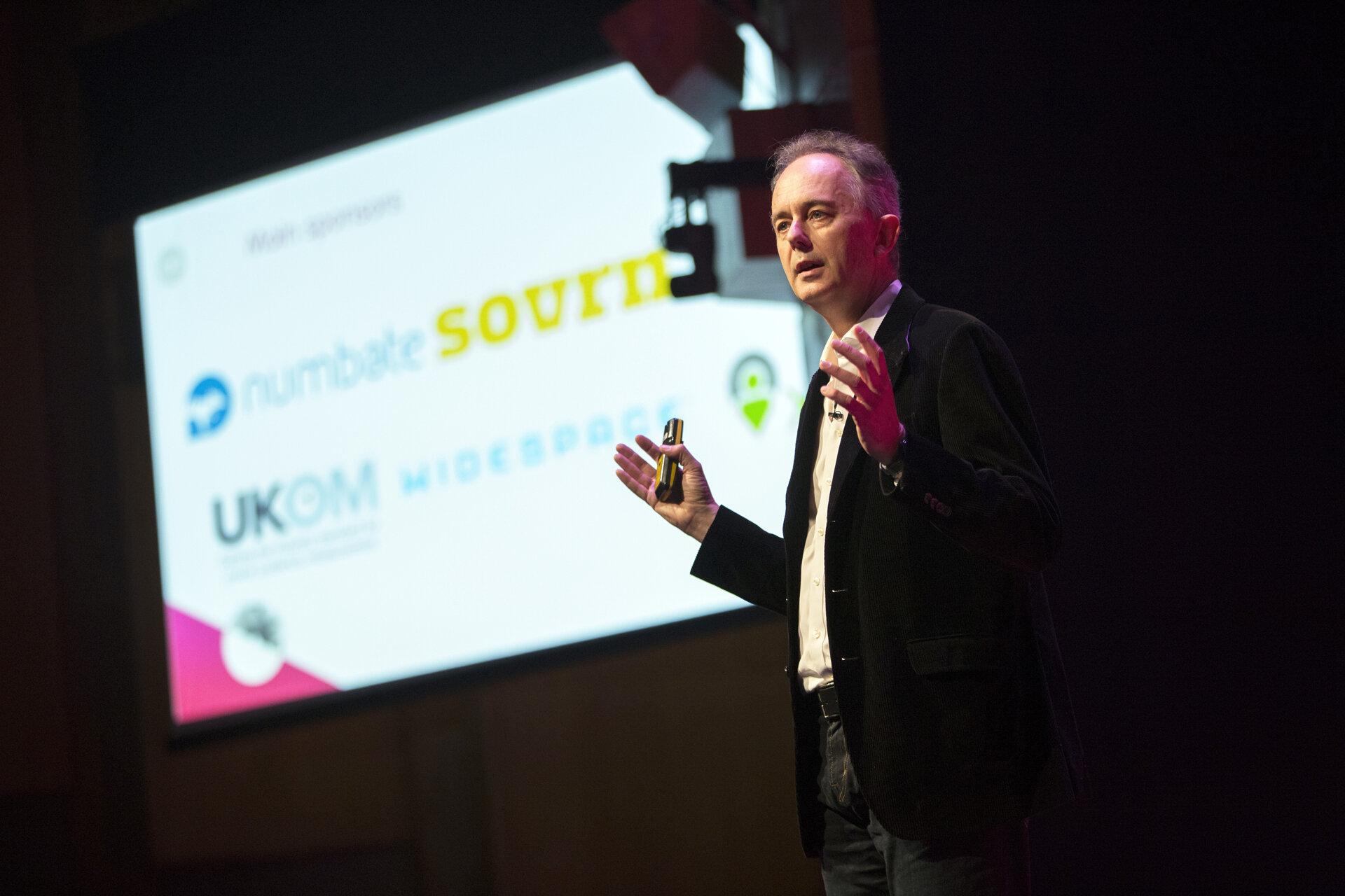 A man delivering a passionate speech on a stage, emphasizing the importance of emotional marketing and avoiding dull advertising, in front of a large screen.