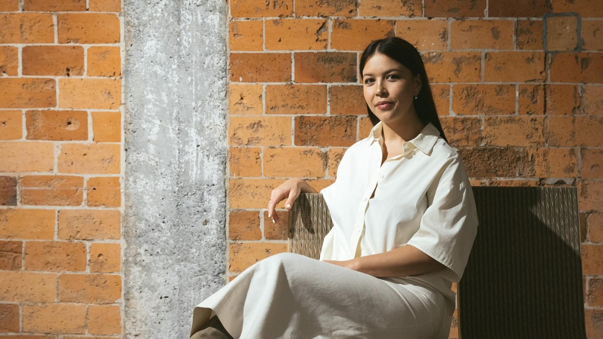 A fashionable woman in a white dress sitting in front of a brick wall, representing the unique and luxury style of the Yu Mei fashion brand