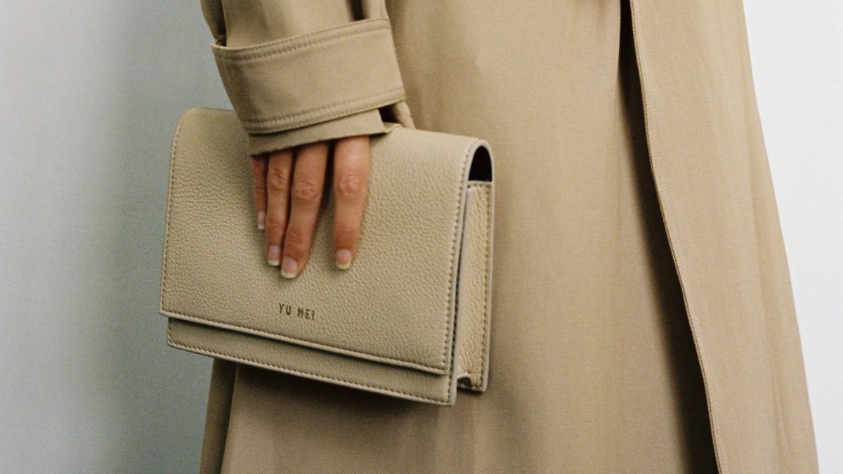 A woman in a trench coat is holding a beige purse.