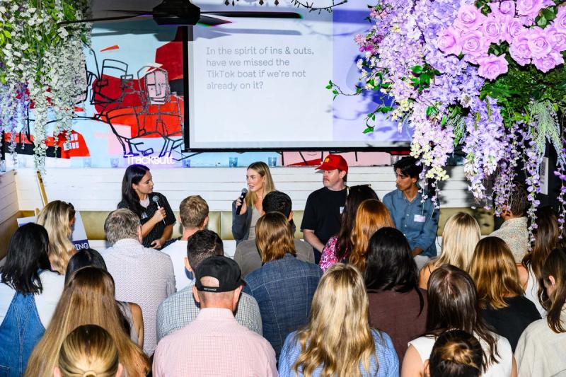 group of panelists in front of an engaged crowd, with purple hanging hydrangeas